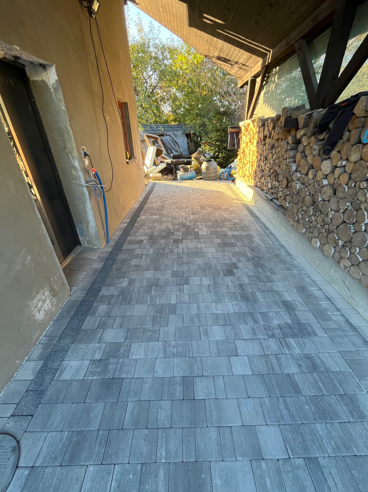 Ułożona szara kostka brukowa na podjeździe, widok z perspektywy wjazdu, obok ściana budynku z oknem i stosem drewna opałowego.