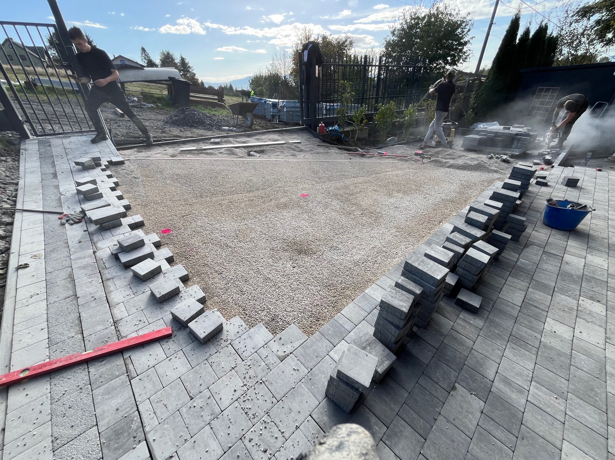 Montaż szarej kostki brukowej na przygotowanym podłożu z jasnego kruszywa, widoczni pracownicy z narzędziami podczas cięcia kostki, ułożona część nawierzchni z poziomnicą, stosy kostki do ułożenia.