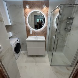 DAMIAN KAPUSZ SPEŁNIAMY WASZE MARZENIA D&M - Nowoczesna łazienka z kabiną prysznicową, umywalką z szafką i pralką, wykończona płytkami imitującymi marmur i drewnianą mozaiką za okrągłym lustrem z podświetleniem LED.