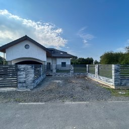 DAMIAN KAPUSZ SPEŁNIAMY WASZE MARZENIA D&M - Nowoczesny dom jednorodzinny z szarą elewacją i grafitowym dachem, otoczony ogrodzeniem z betonowych bloczków imitujących kamień i metalowych przęseł, widok z zewnątrz na podjazd wysypany żwirem.