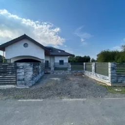 Nowoczesny dom jednorodzinny z szarą elewacją i grafitowym dachem, otoczony ogrodzeniem z betonowych bloczków imitujących kamień i metalowych przęseł, widok z zewnątrz na podjazd wysypany żwirem.