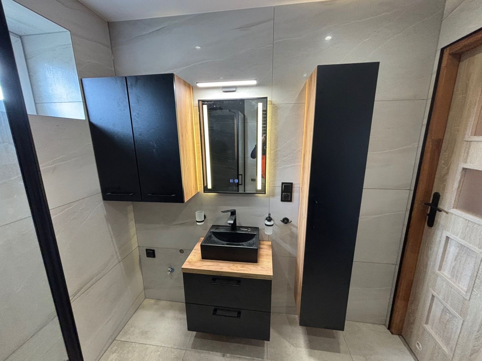 Nowoczesna łazienka z czarną umywalką na drewnianym blacie, podświetlanym lustrem i szafkami w kolorze czarnym, wykończona płytkami imitującymi kamień.