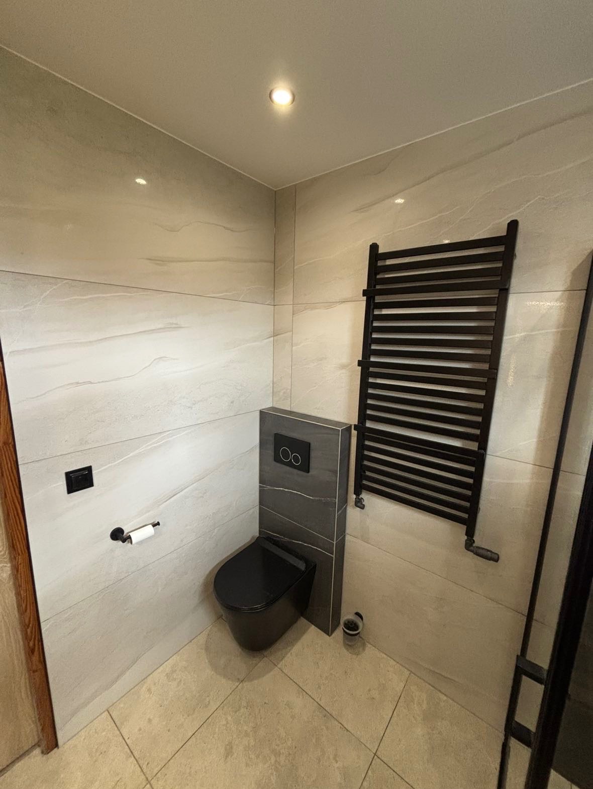 Nowoczesna łazienka z beżowymi płytkami imitującymi marmur, czarną wiszącą toaletą i grzejnikiem drabinkowym w kolorze czarnym. Widoczny również czarny przycisk spłuczki podtynkowy i uchwyt...