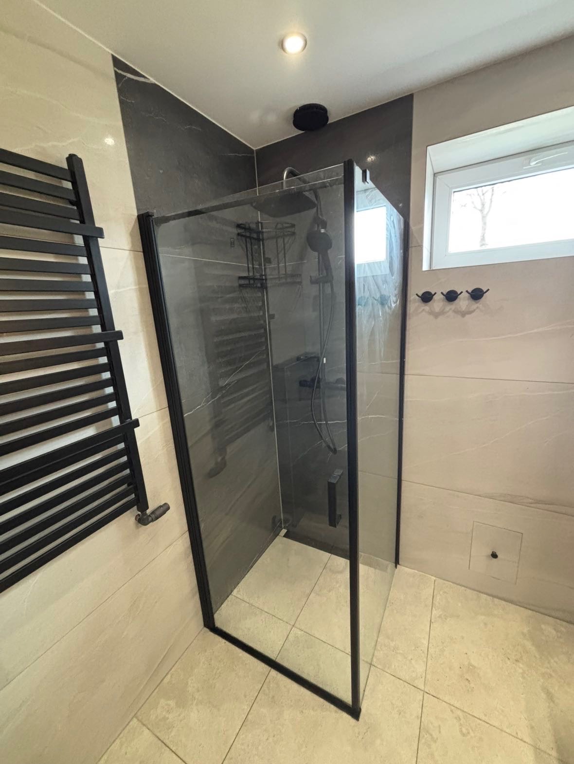 Nowoczesna kabina prysznicowa z czarnymi ramami, zamontowana w narożniku łazienki z beżowymi i szarymi płytkami, obok grzejnik drabinkowy w kolorze czarnym i okno z trzema haczykami pod spodem.