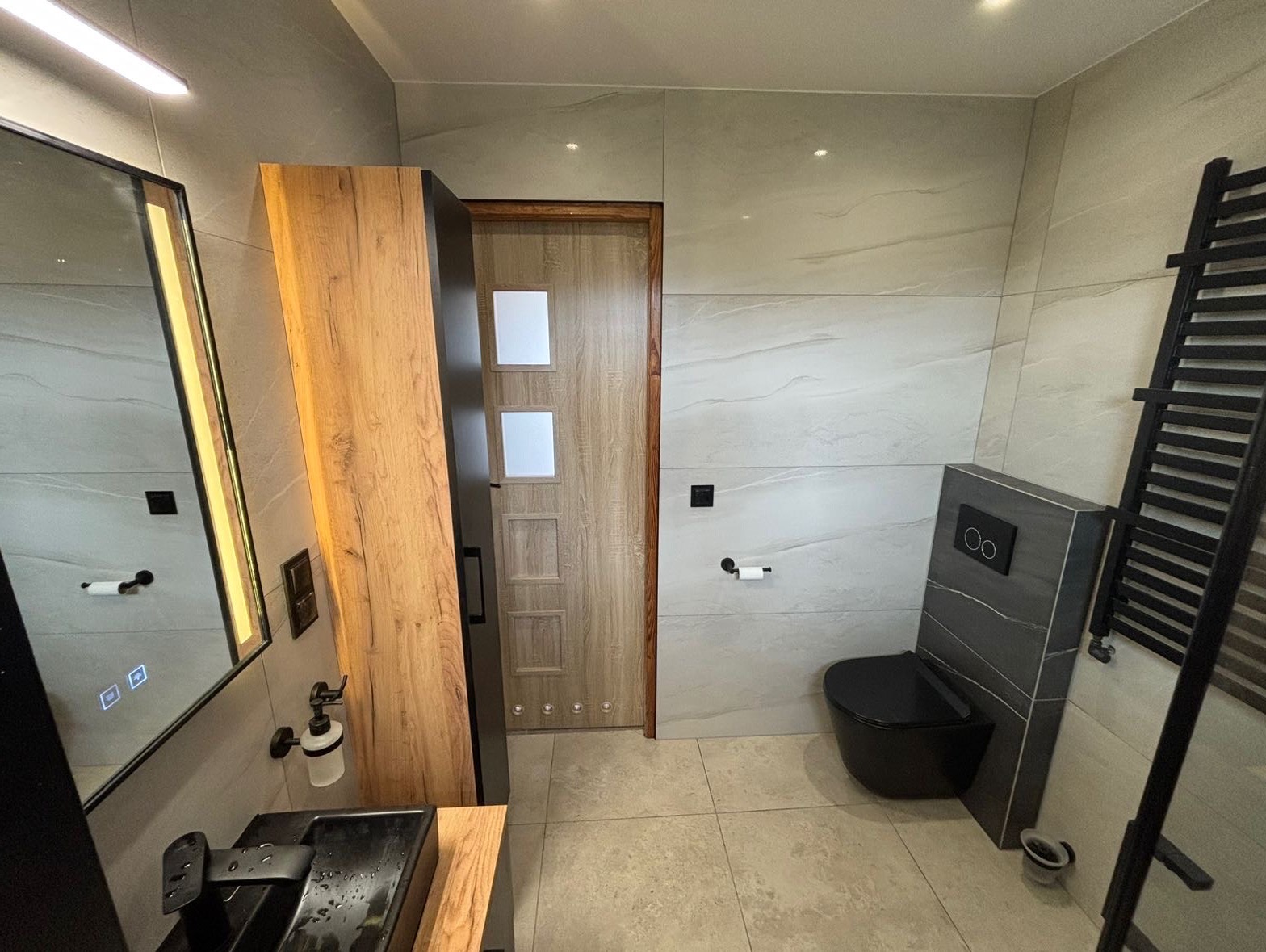 Nowoczesna łazienka z szarymi płytkami imitującymi marmur, czarną armaturą i drewnianymi elementami wykończenia, w tym szafką z lustrem z podświetleniem LED i drzwiami z przeszkleniami.