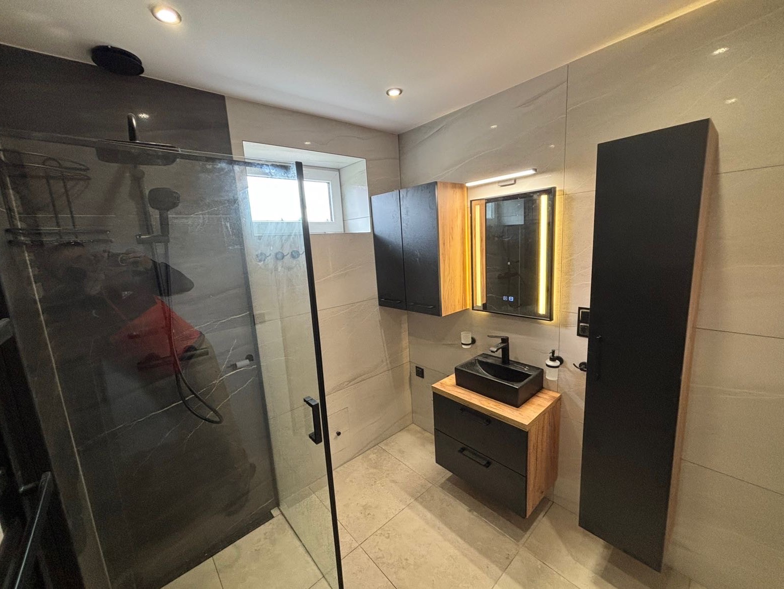Nowoczesna łazienka z kabiną prysznicową typu walk-in, umywalką nablatową w kolorze czarnym, szafką pod umywalkową z drewnianym blatem, podświetlanym lustrem i płytkami imitującymi marmur