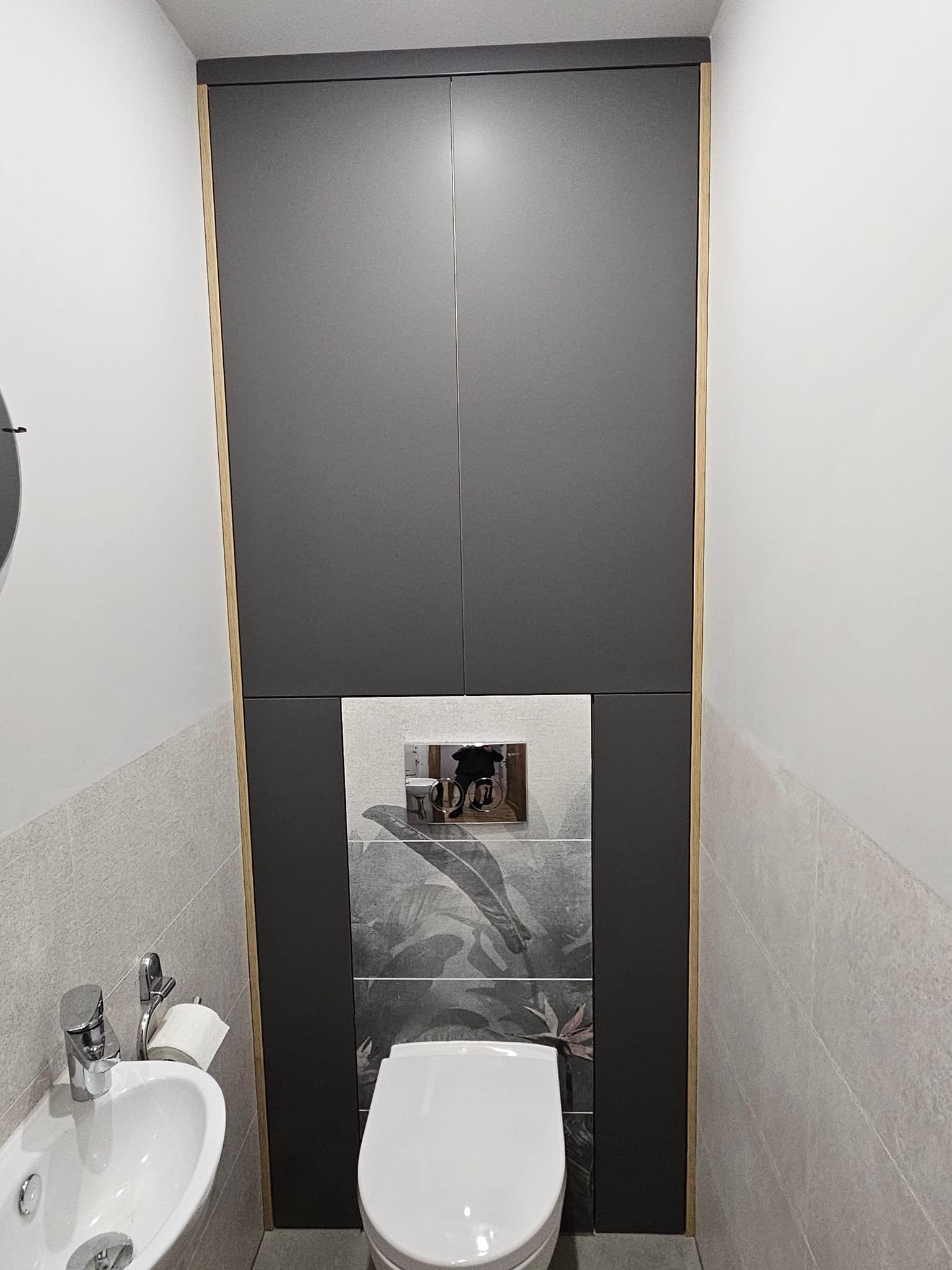 Nowoczesna toaleta w szarościach z zabudową nad stelażem WC, akcentem roślinnym na płytkach i minimalistyczną umywalką.