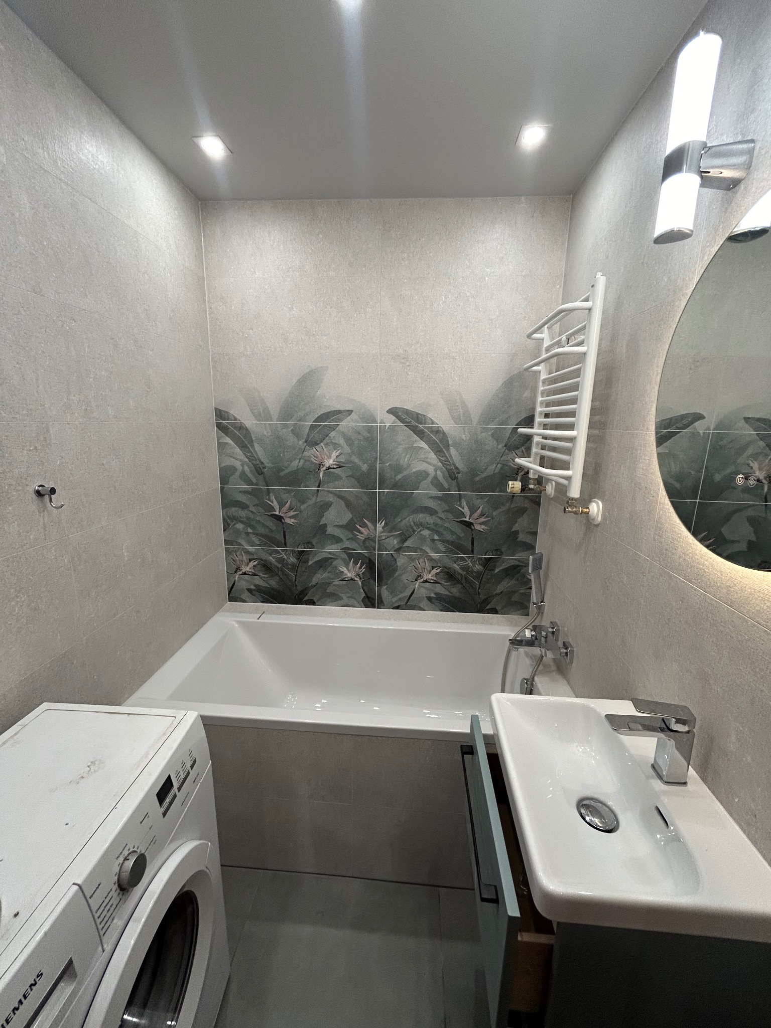 Nowoczesna łazienka z płytkami imitującymi kamień, akcentem w postaci płytek z motywem tropikalnych liści nad wanną, okrągłym lustrem z podświetleniem LED, białą umywalką z szafką i pralką Siemens.