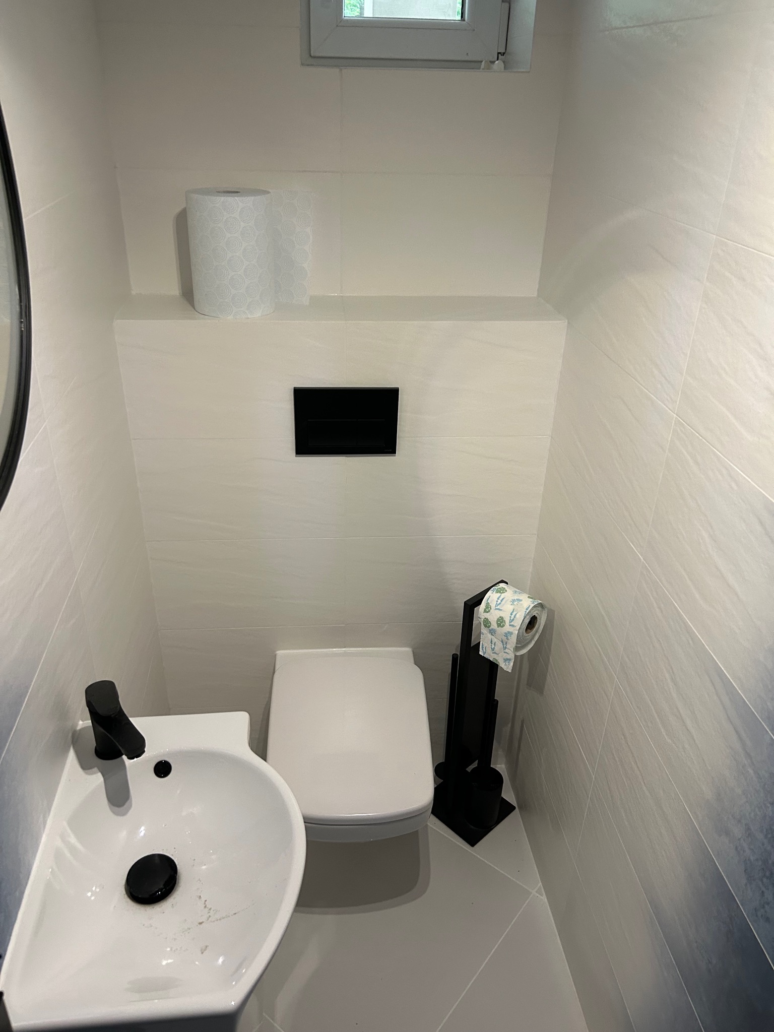 Mała, nowoczesna toaleta z umywalką narożną, białą ceramiką i płytkami imitującymi kamień w odcieniach bieli i szarości, z czarnymi akcentami w postaci baterii i uchwytu na papier toaletowy.