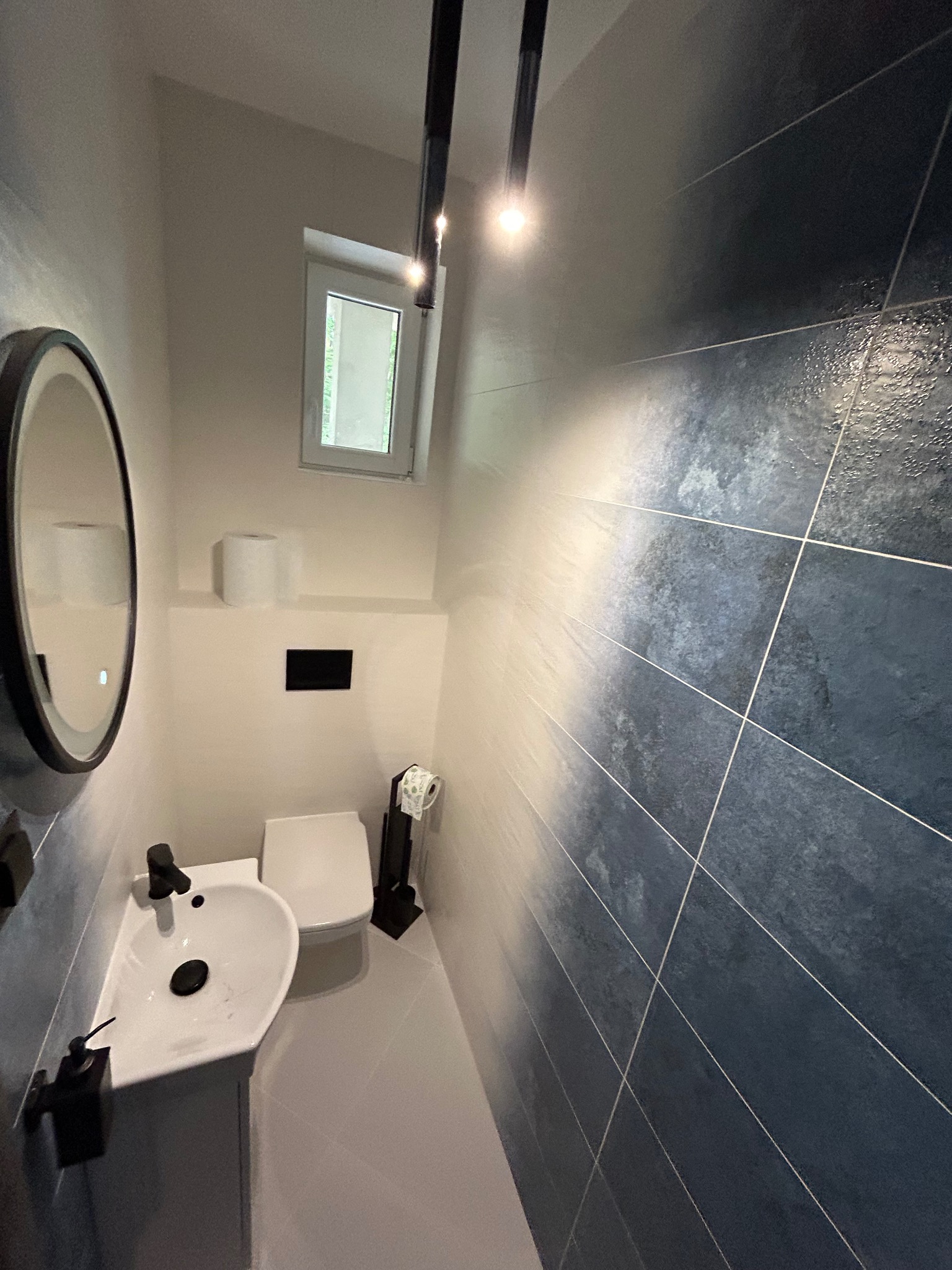 Nowoczesna toaleta z umywalką narożną, podwieszaną miską WC i ciemnymi płytkami imitującymi kamień na ścianie, oświetlona lampami sufitowymi.