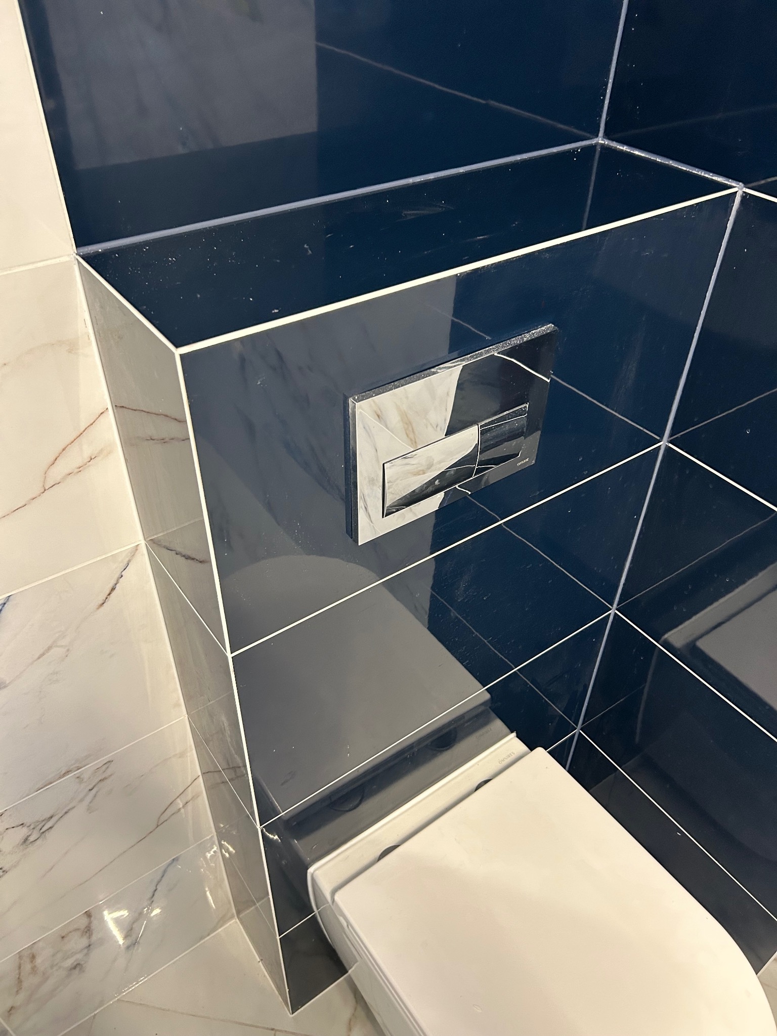 Detale wykończenia łazienki: połączenie białych płytek imitujących marmur z ciemnogranatowymi, błyszczącymi płytkami ceramicznymi, widoczny chromowany przycisk spłuczki, fragment toalety.