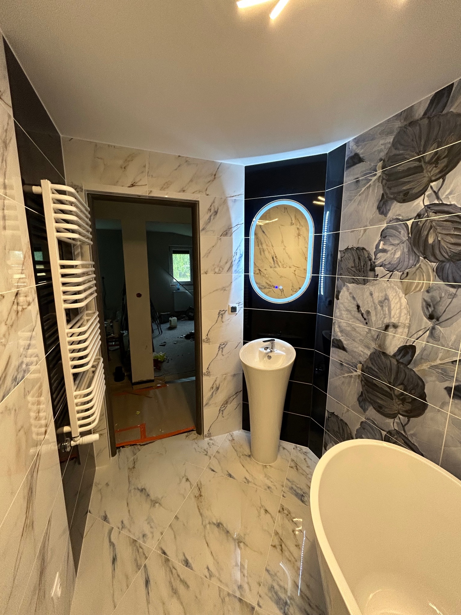 Nowoczesna łazienka z marmurowymi płytkami, wolnostojącą umywalką na postumencie, podświetlanym lustrem owalnym i grzejnikiem drabinkowym; widok na pomieszczenie w trakcie remontu przez otwarte drzwi.