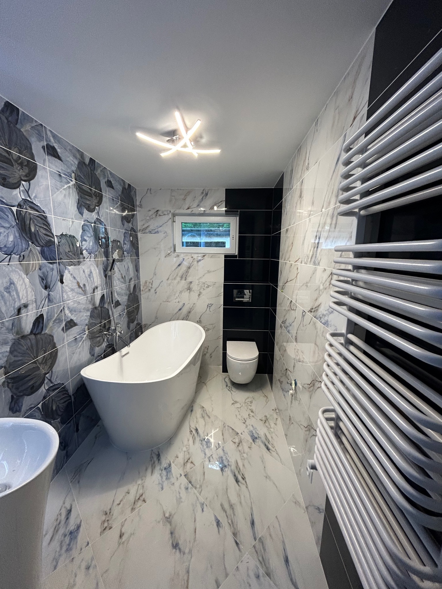 Nowoczesna łazienka z wolnostojącą wanną, marmurowymi płytkami z motywem liści, grzejnikiem drabinkowym i minimalistycznym oświetleniem sufitowym.