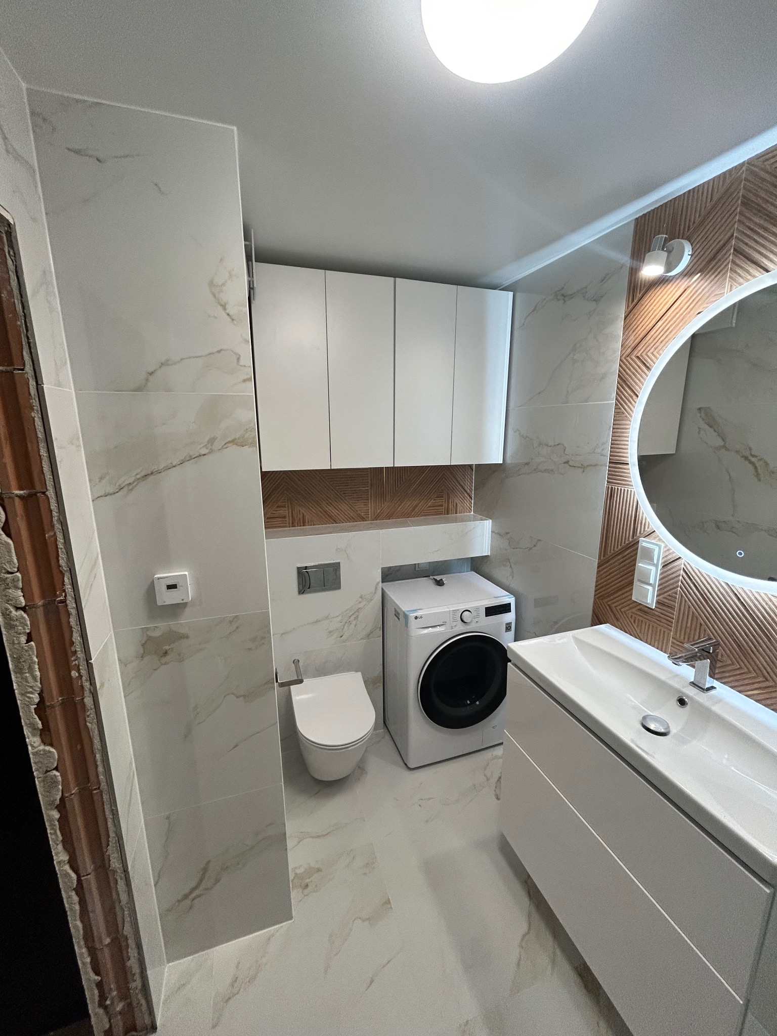 Nowoczesna łazienka z marmurowymi płytkami, podświetlanym okrągłym lustrem, wiszącą szafką, pralką i wiszącą toaletą.