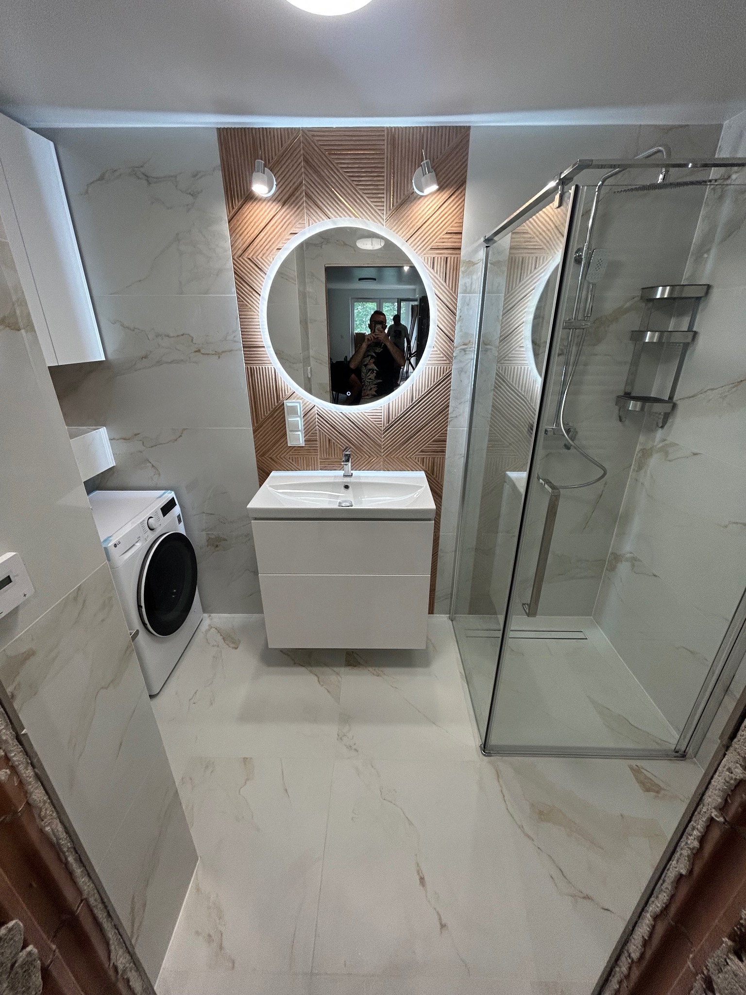 Nowoczesna łazienka z kabiną prysznicową, umywalką z szafką i pralką, wykończona płytkami imitującymi marmur i drewnianą mozaiką za okrągłym lustrem z podświetleniem LED.