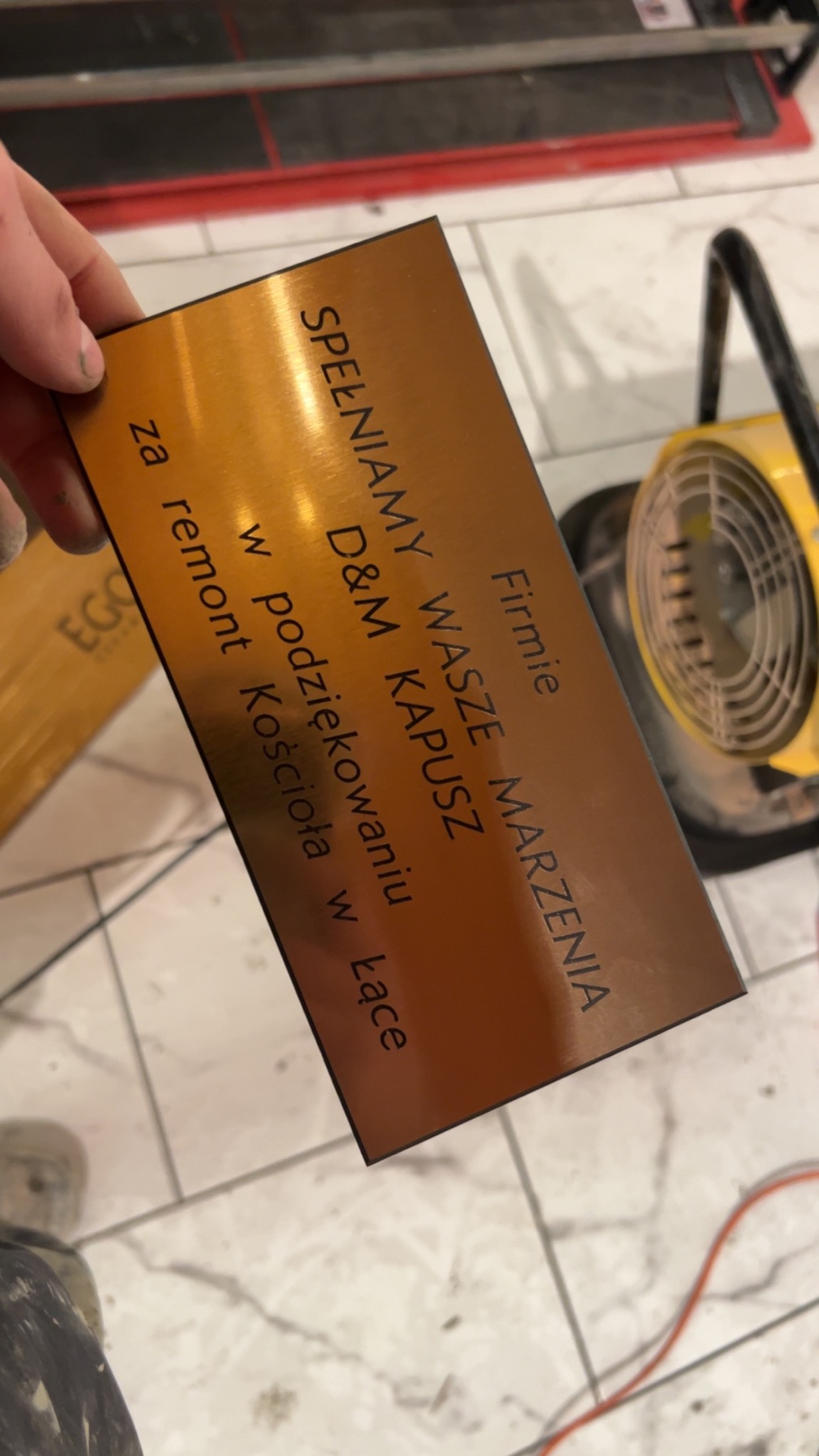 Brązowa tabliczka z grawerowanym napisem 'Firmie D&M KAPUSZ SPEŁNIAMY WASZE MARZENIA w podziękowaniu za remont Kościoła w Łące' trzymana w dłoni na tle płytek podłogowych i narzędzi glazurniczych.