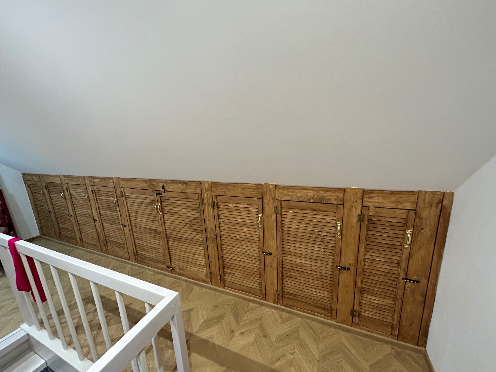 Długi rząd wbudowanych, drewnianych szafek z żaluzjowymi drzwiczkami pod skosem dachu na poddaszu, widoczny fragment białej balustrady schodów.