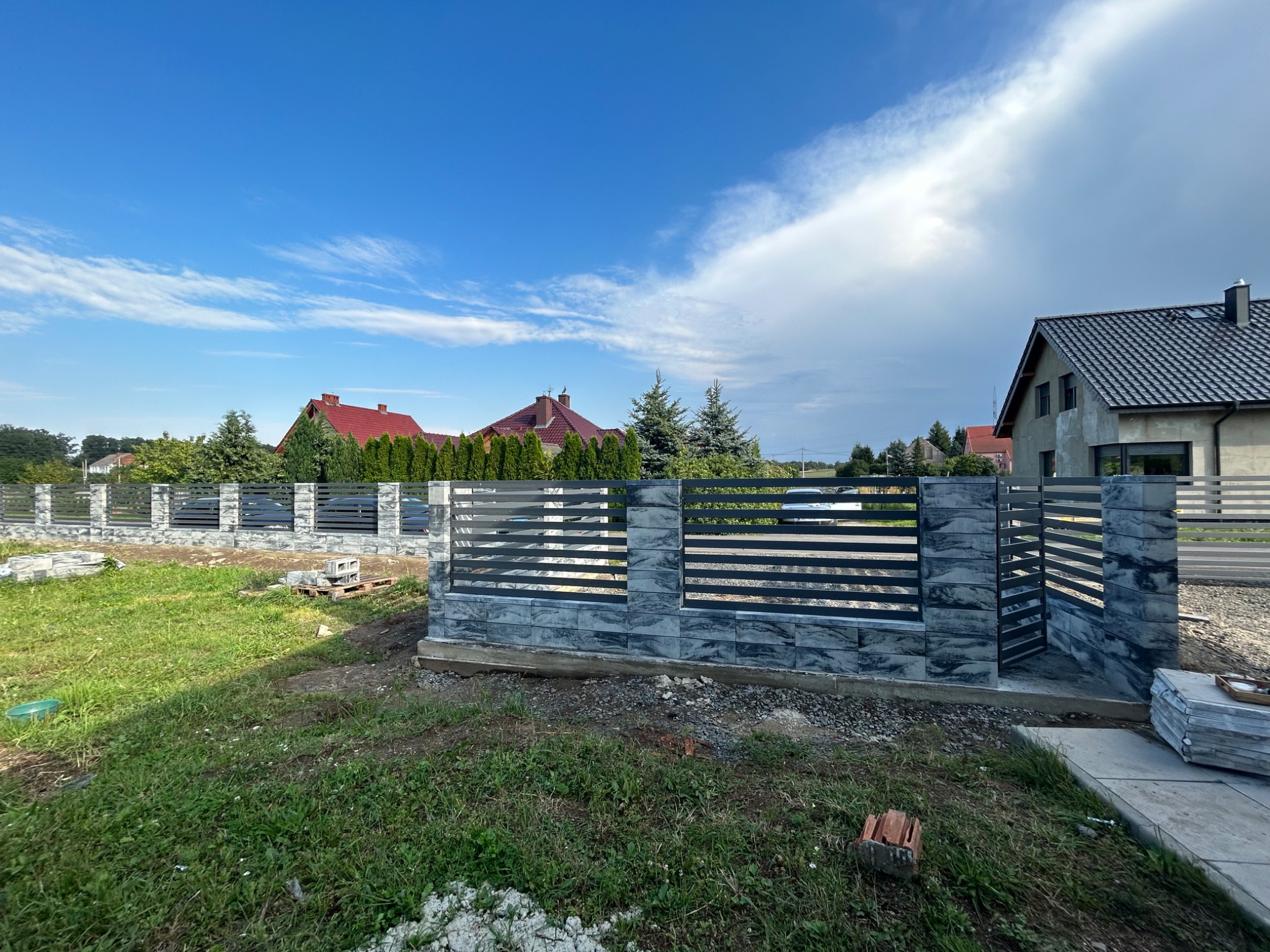 Nowoczesne ogrodzenie panelowe z elementami imitującymi kamień, w trakcie montażu, widoczne niezakończone prace na trawniku.