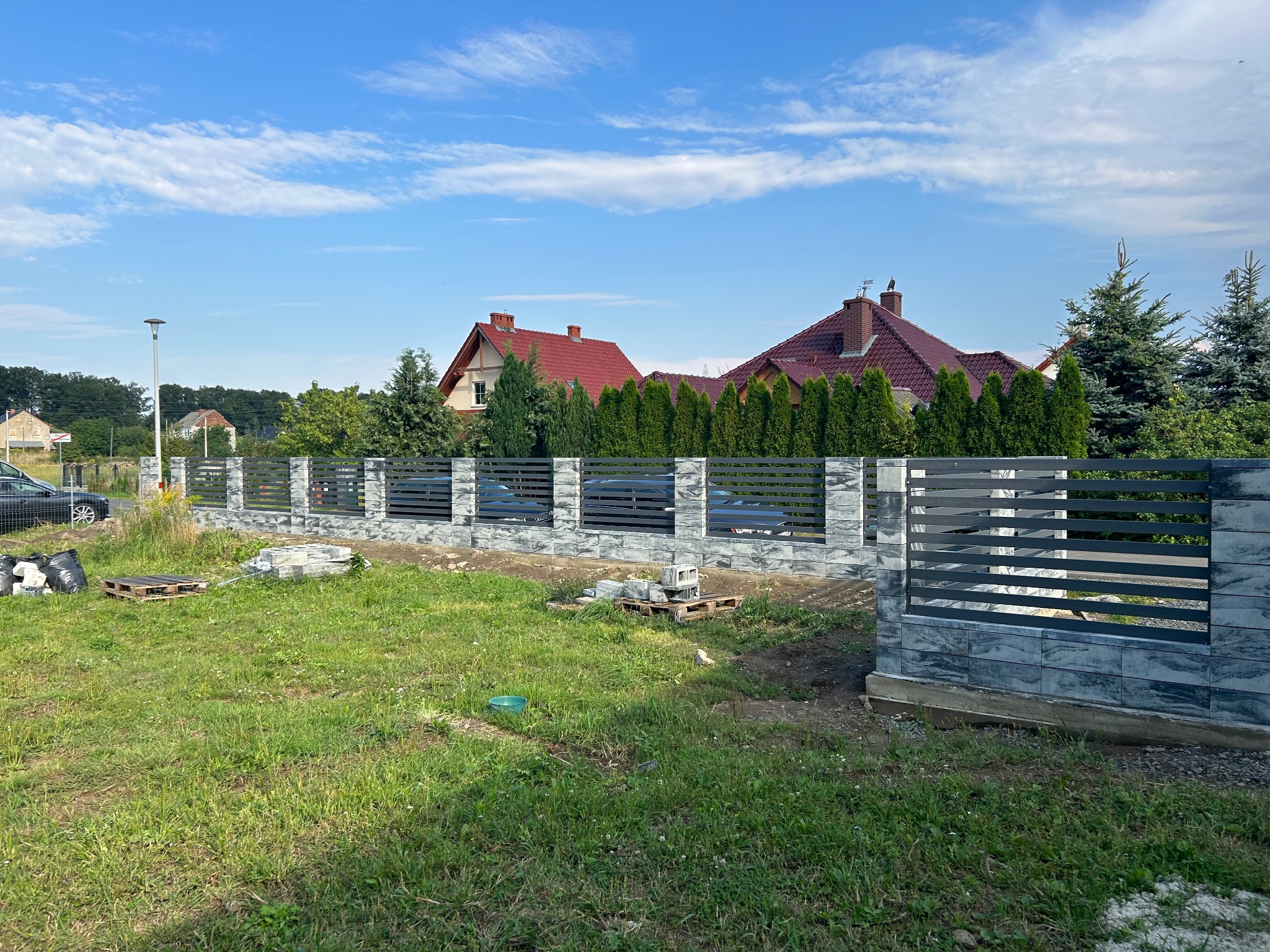 Montaż ogrodzenia panelowego z elementami imitującymi kamień na tle domów jednorodzinnych w słoneczny dzień.