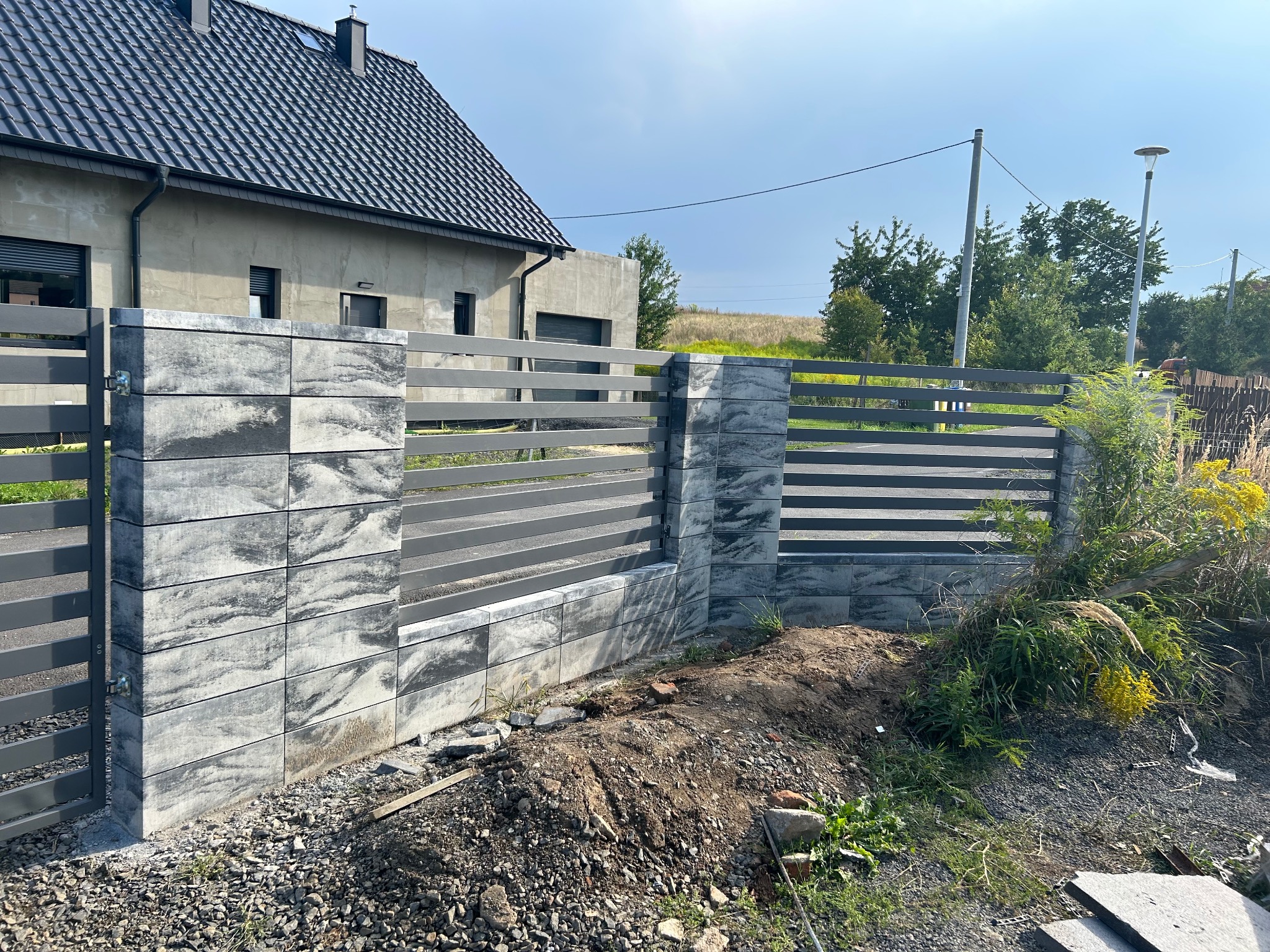 Nowoczesne ogrodzenie panelowe z elementami betonowymi o wzorze marmurkowym, w trakcie montażu; widoczne niedokończone prace ziemne i gruz.