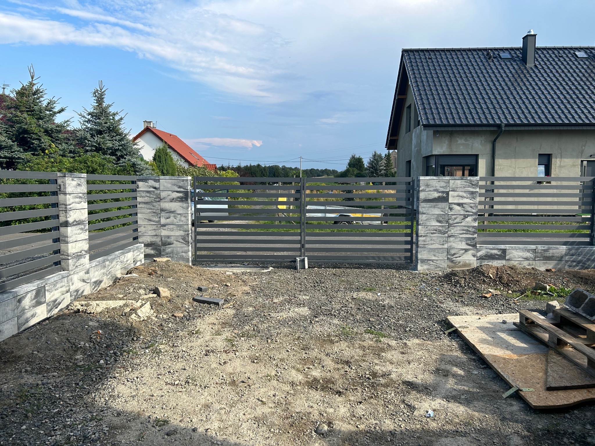 Nowoczesne ogrodzenie panelowe z betonowymi słupkami i bramą wjazdową na tle domu w trakcie budowy, teren niezagospodarowany.