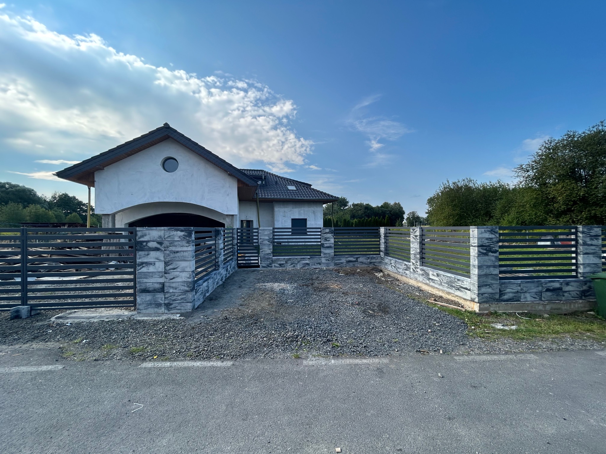 Nowoczesny dom jednorodzinny z szarą elewacją i grafitowym dachem, otoczony ogrodzeniem z betonowych bloczków imitujących kamień i metalowych przęseł, widok z zewnątrz na podjazd wysypany żwirem.