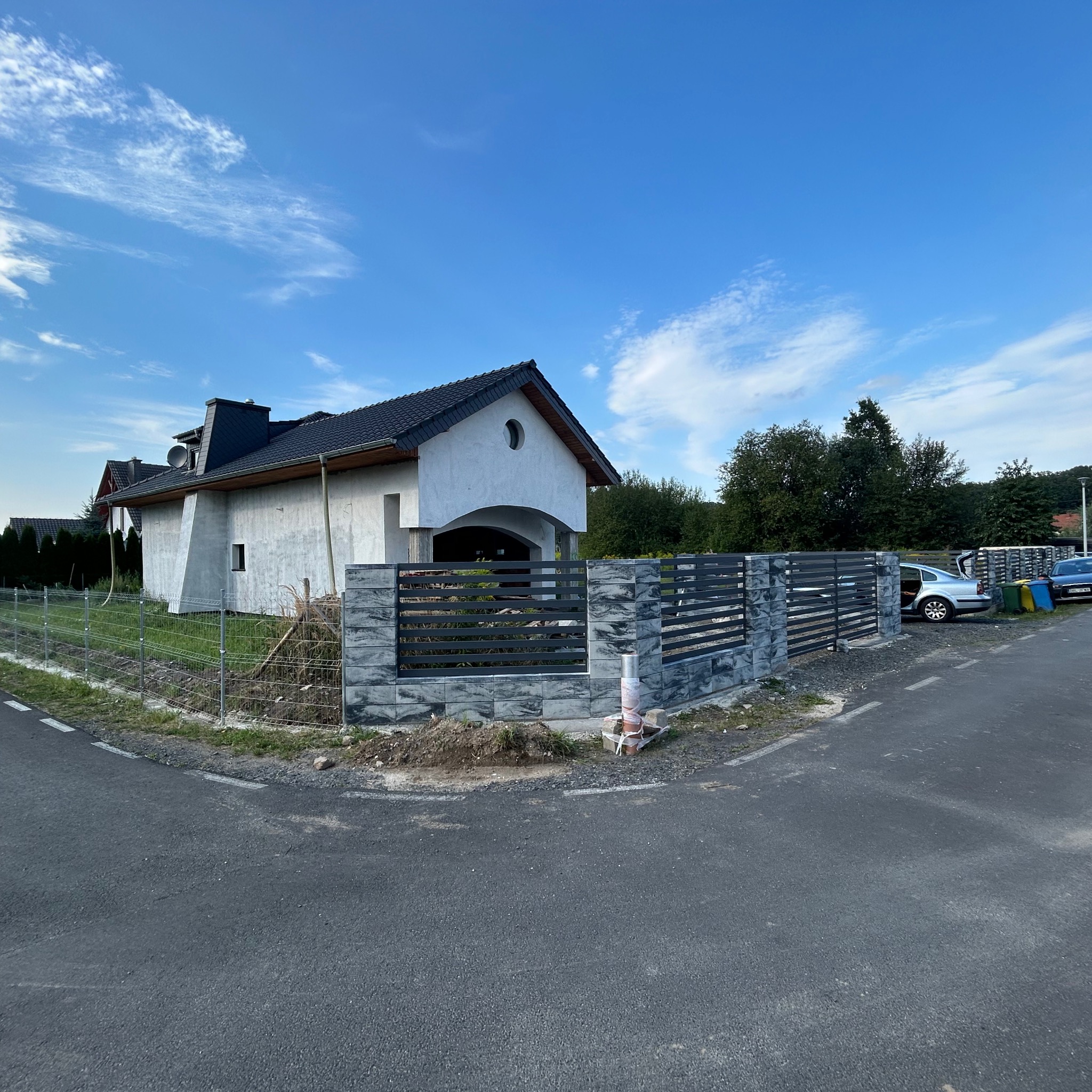 Nowoczesny dom jednorodzinny z częściowo ukończonym ogrodzeniem z szarego kamienia i metalowych przęseł, widoczne tymczasowe zabezpieczenia budowy i materiały budowlane.