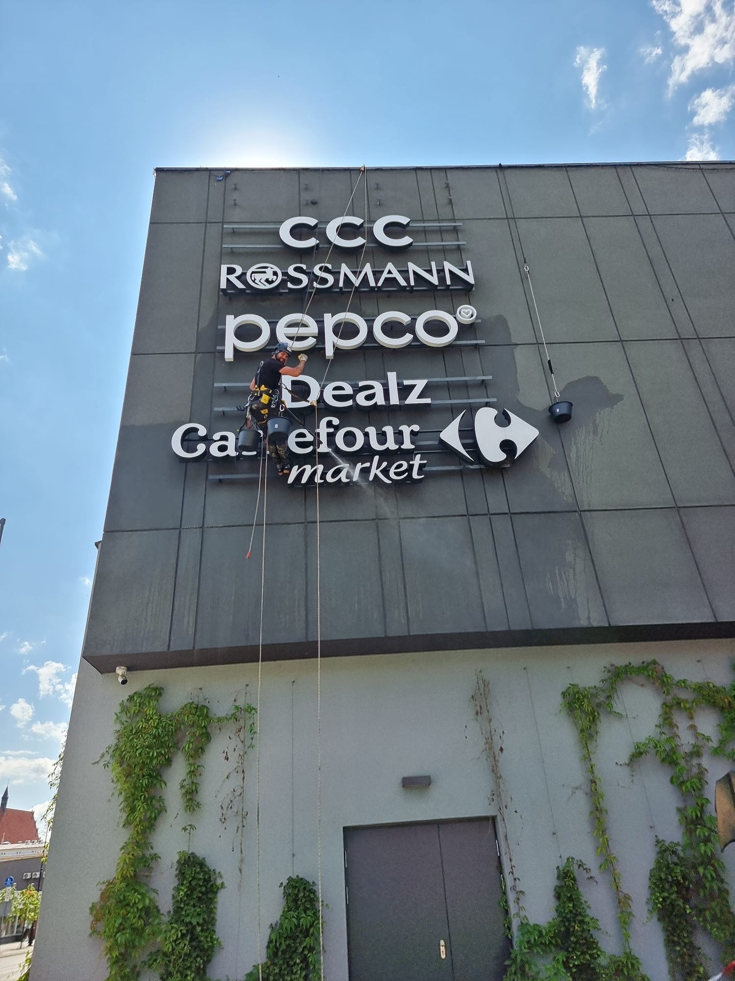Pracownik na linach czyści elewację budynku z logotypami sklepów, widoczne ślady mycia. Budynek z szarą elewacją i zielonymi pnączami. Błękitne niebo w tle.