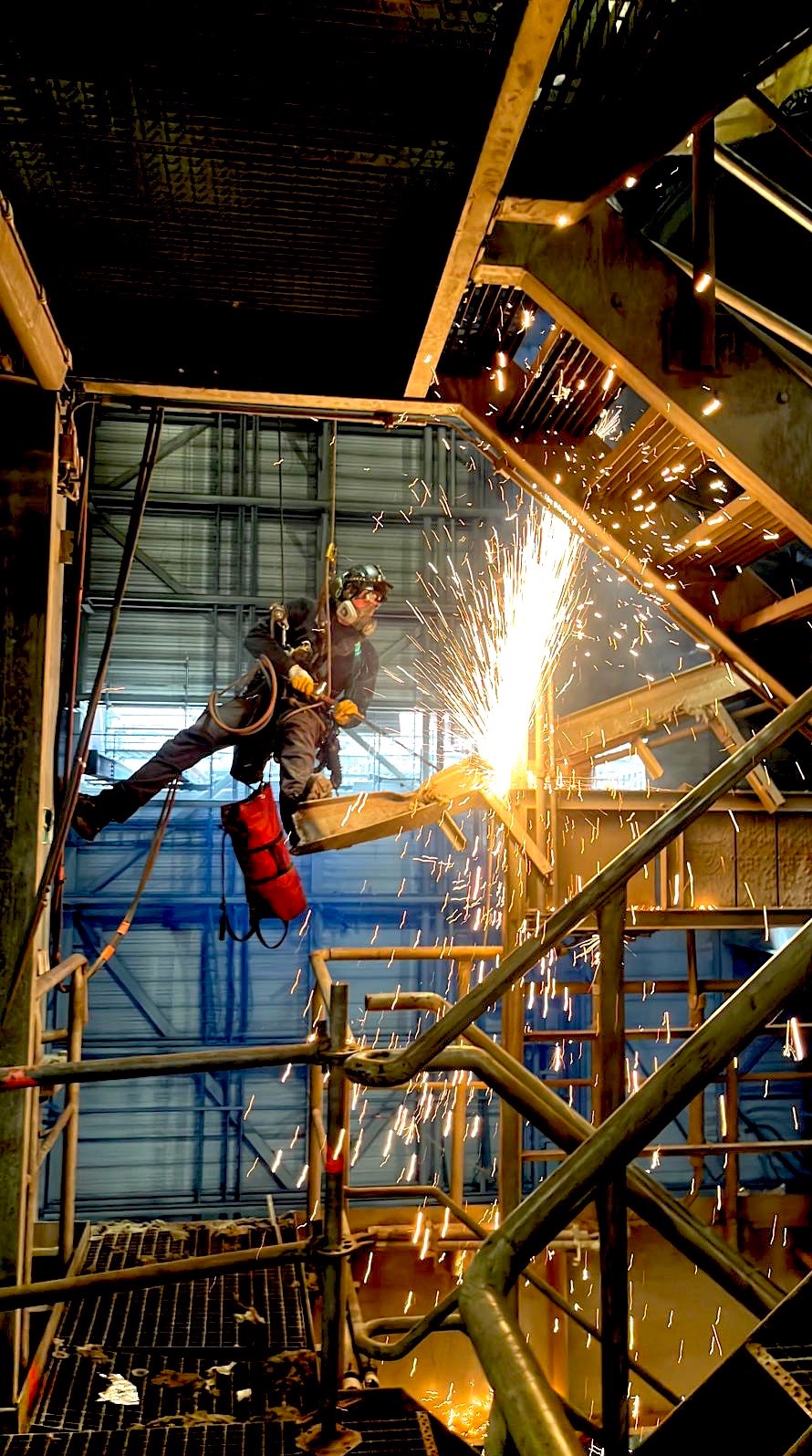 Pracownik w uprzęży, w kasku i masce spawalniczej, szlifuje metalową konstrukcję na wysokości, iskry rozbryzgują się w powietrzu. Widok z dołu.