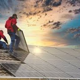 Raccoon services - Dwóch monterów w czerwonych kurtkach instaluje panele słoneczne na dachu domu, widoczne narzędzia i części instalacji, malownicze chmury na niebie w tle.