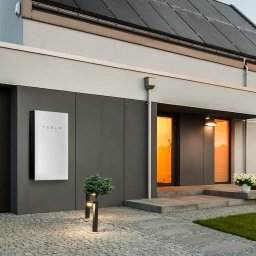 Raccoon services - Nowoczesny dom z panelami fotowoltaicznymi na dachu i zewnętrznym akumulatorem energii Tesla Powerwall zamontowanym na ścianie, oświetlonymi ścieżkami i zielonym trawnikiem.