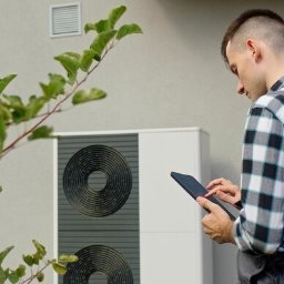 Raccoon services - Instalator w kombinezonie w kratę sprawdza dane na tablecie obok białej pompy ciepła z dwoma okrągłymi wlotami powietrza na tle jasnej ściany budynku, z zielonymi liśćmi krzewu po lewej stronie kadru.