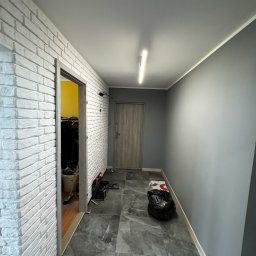 Piotr Car PIOTR SKOTNICKI - Korytarz po remoncie: biała ściana z cegły, szara podłoga z płytek imitujących marmur, szare ściany. Widoczne narzędzia i materiały budowlane.