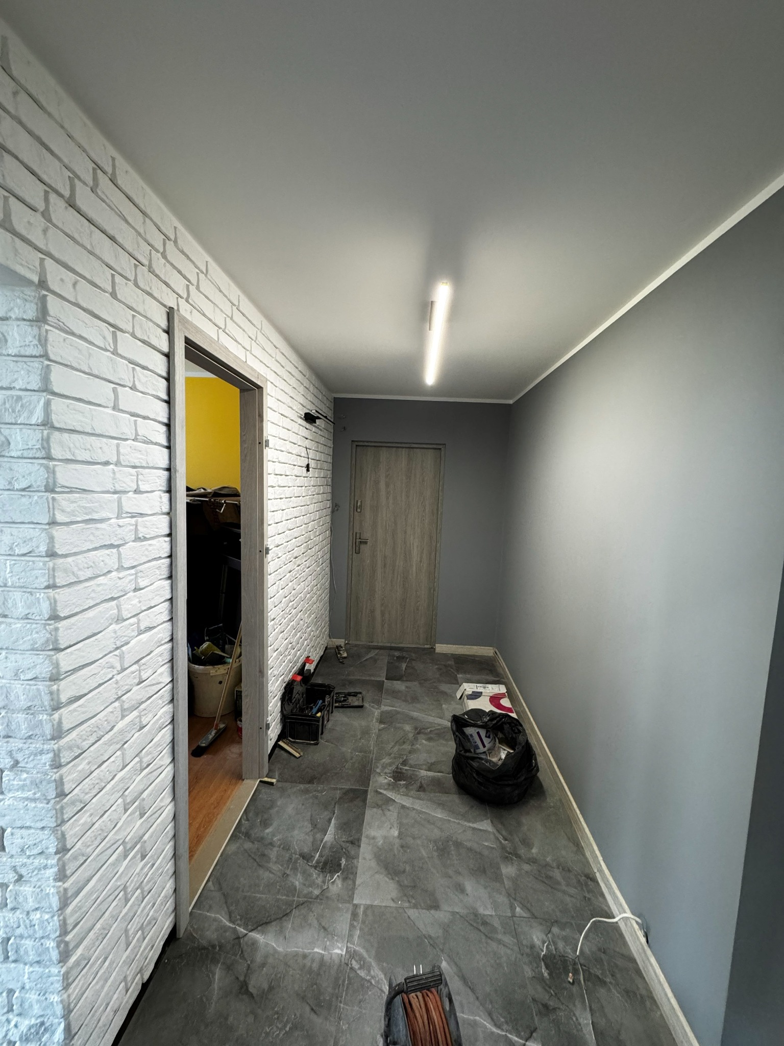 Korytarz po remoncie: biała ściana z cegły, szara podłoga z płytek imitujących marmur, szare ściany. Widoczne narzędzia i materiały budowlane.