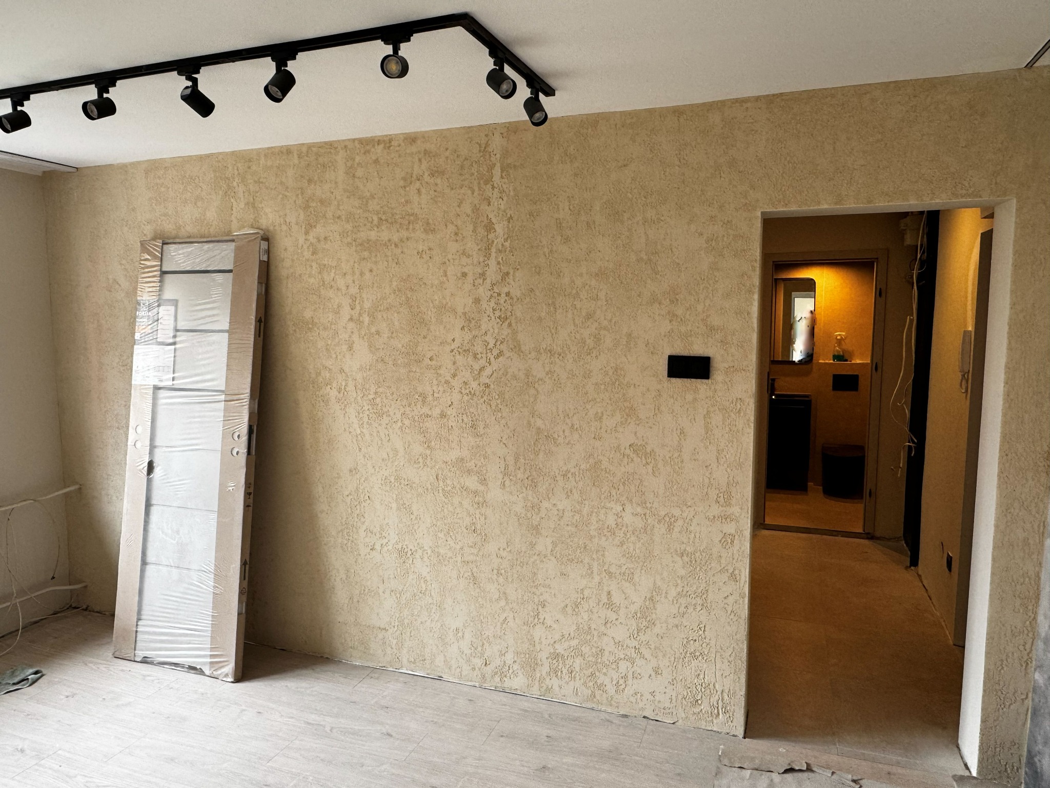 Wykończone ściany z fakturą w odcieniach beżu, oświetlenie sufitowe punktowe, drzwi wewnętrzne w folii ochronnej i widok na łazienkę. Elementy wykończenia wnętrz.