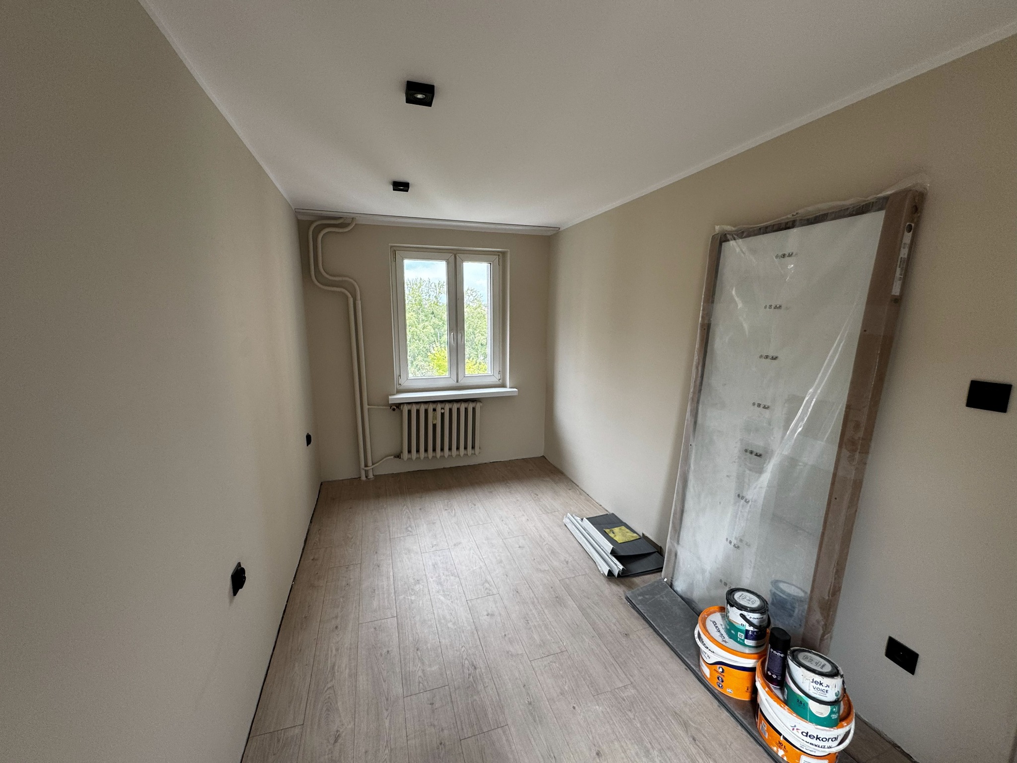 Pokój po remoncie: świeżo pomalowane ściany w odcieniach beżu, jasna podłoga, okno z widokiem na zieleń, drzwi w folii i farby Dekoral. Widoczne gniazdka elektryczne.