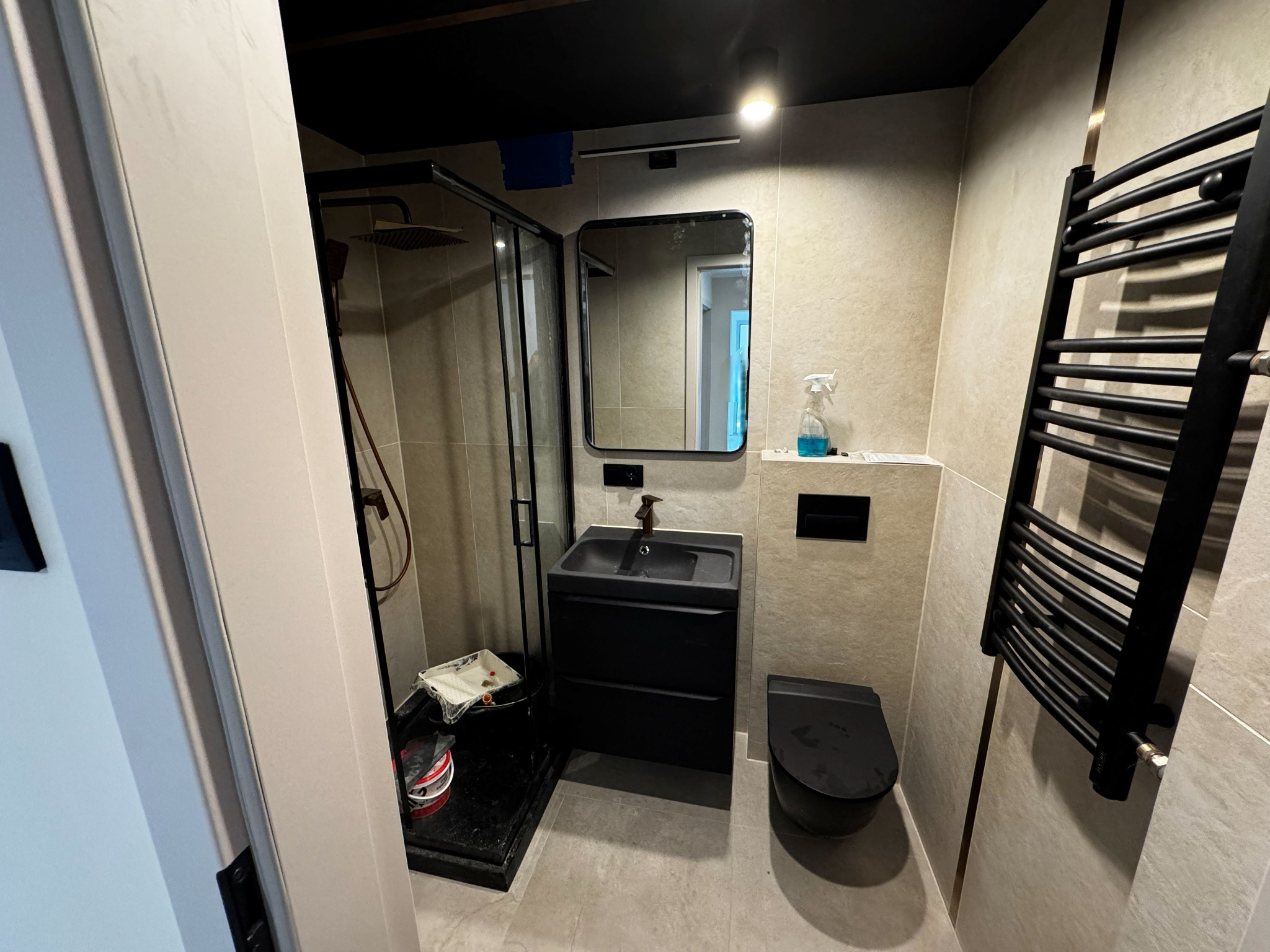 Nowoczesna łazienka w odcieniach beżu i czerni: kabina prysznicowa typu walk-in, umywalka z szafką, wisząca toaleta i grzejnik drabinkowy. Minimalistyczny design.