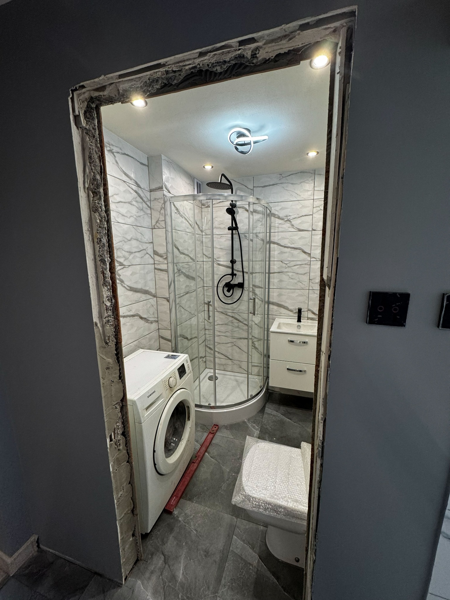 Remont łazienki w trakcie: kabina prysznicowa, umywalka z szafką, pralka i toaleta w folii ochronnej. Widoczne niedokończone obramowanie drzwi.