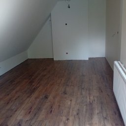 FIRMA WYKOŃCZENIOWO BUDOWLANA Krzysztof Pieniak - Podłoga z paneli w wykończonym pokoju na poddaszu. Widoczne białe ściany, grzejnik i instalacja elektryczna. Ukończony remont, ciepłe odcienie brązu.