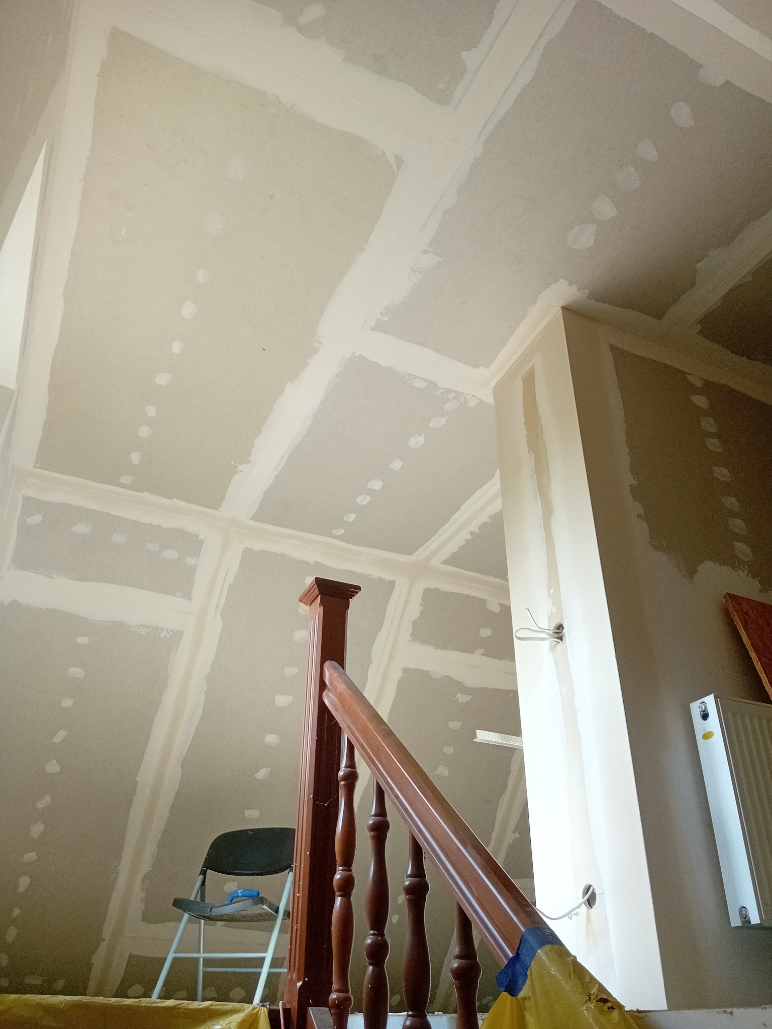 Sufit i ściany po montażu płyt gipsowo-kartonowych, widoczne łączenia i ślady szpachlowania. W tle fragment schodów z drewnianą balustradą i krzesło.