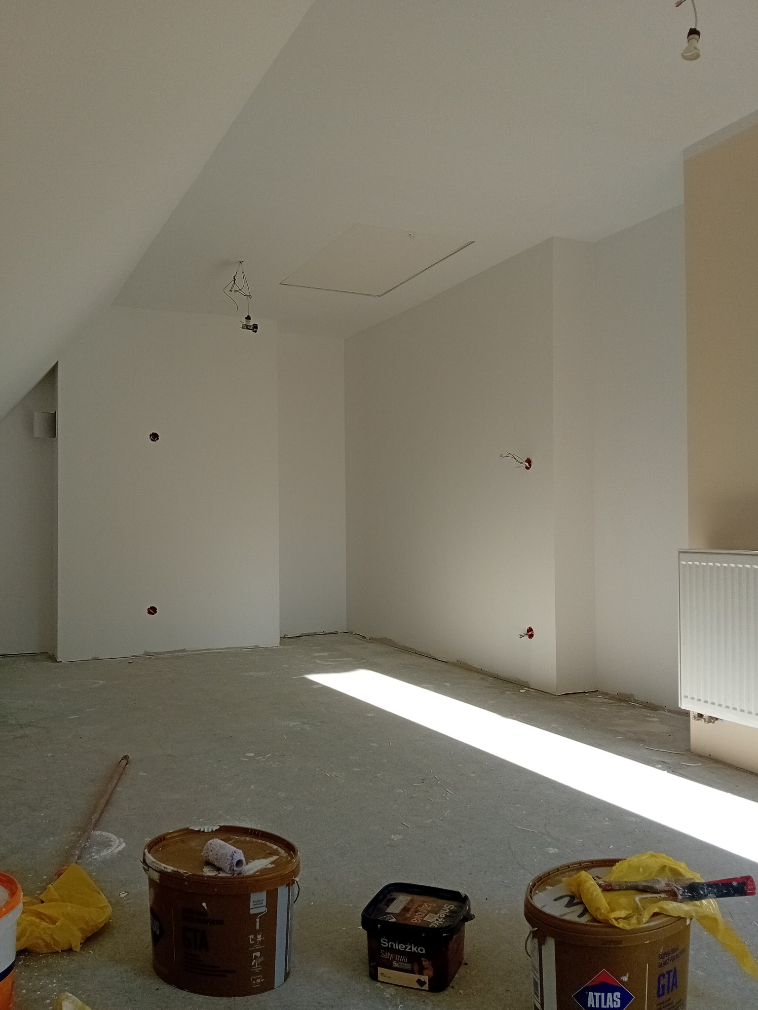 Wnętrze po malowaniu: białe ściany pod skosem, widoczne puszki z farbą Atlas i Śnieżka, wałek malarski. Przygotowane do dalszych prac wykończeniowych.