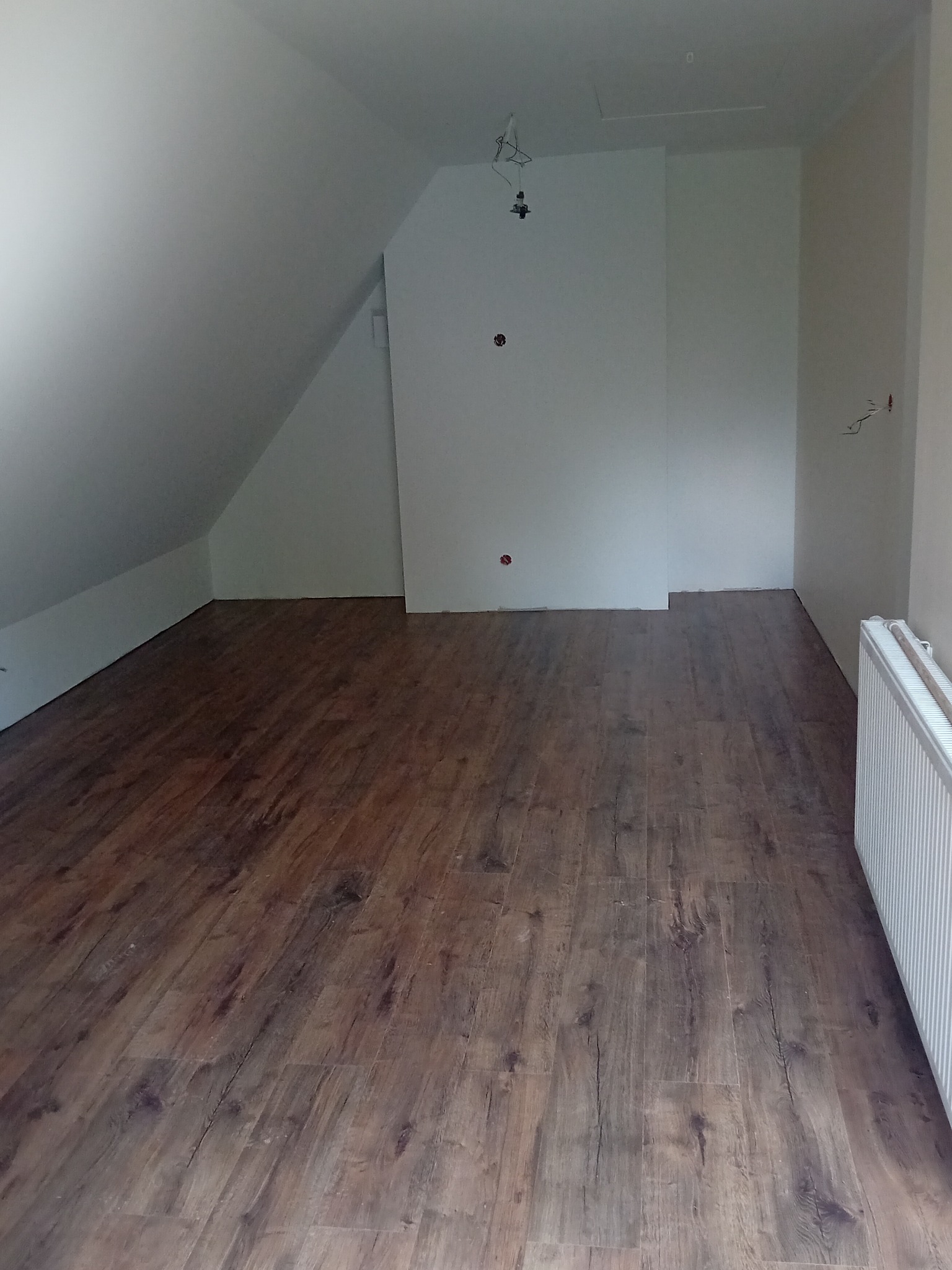 Podłoga z paneli w wykończonym pokoju na poddaszu. Widoczne białe ściany, grzejnik i instalacja elektryczna. Ukończony remont, ciepłe odcienie brązu.