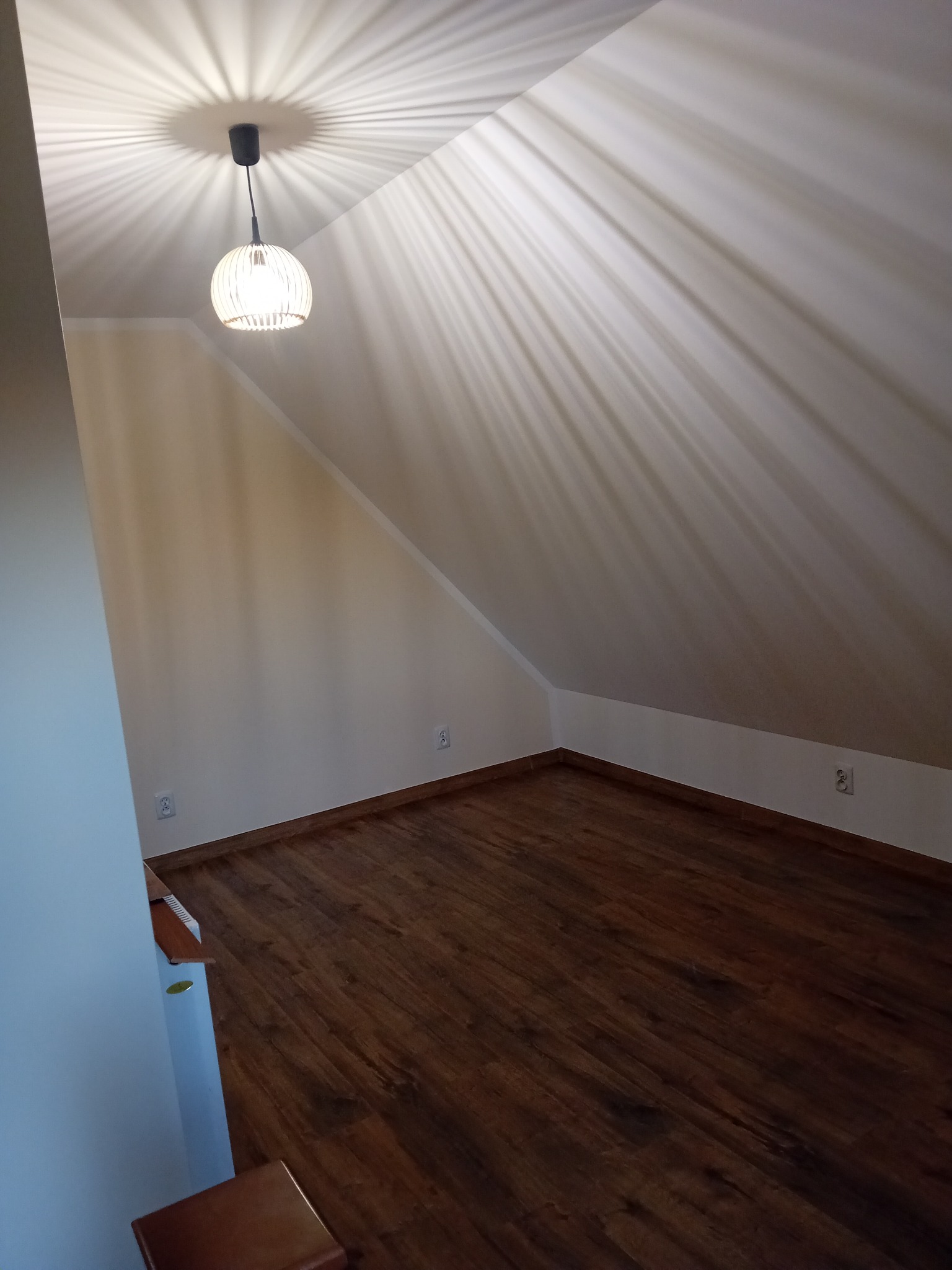 Poddasze po remoncie: drewniana podłoga, białe ściany, nietypowe oświetlenie sufitowe tworzące promienie. Widok z dołu, ukończone gniazdka elektryczne.
