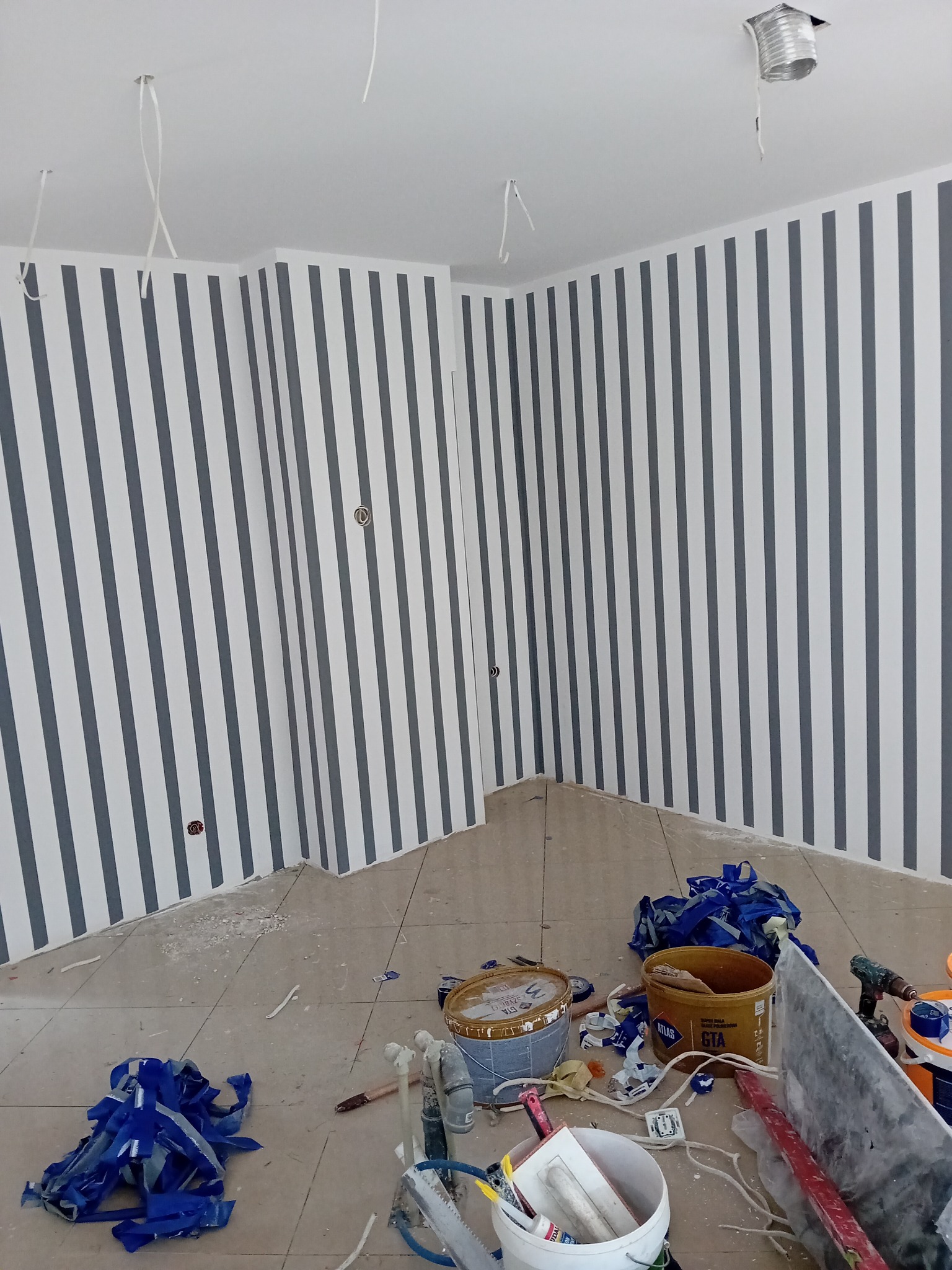 Pomieszczenie po malowaniu ścian w szaro-białe pasy. Na podłodze widoczne narzędzia malarskie, pędzle, wałki i resztki farby. Sufit biały z wystającymi kablami.