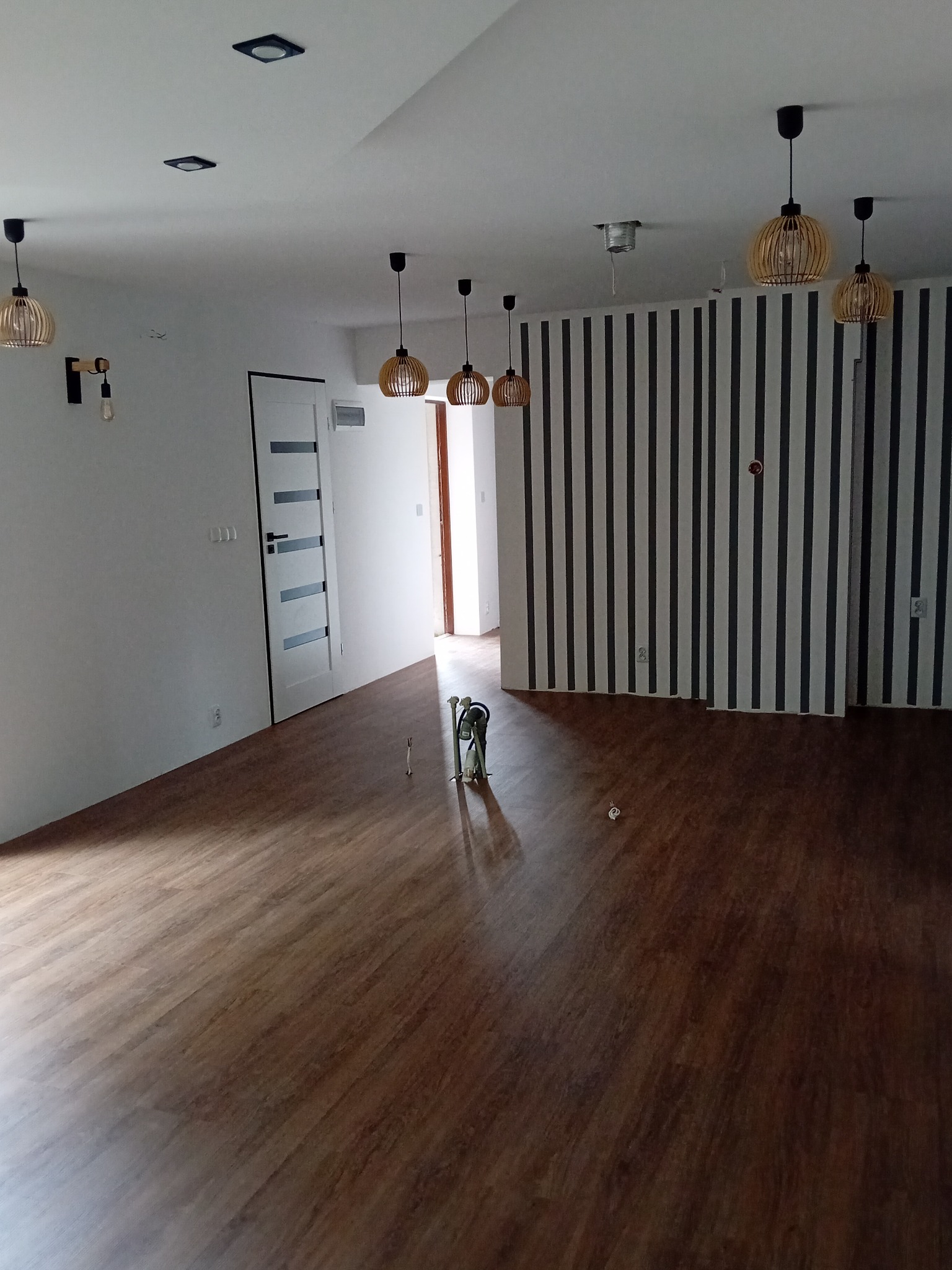 Jasne wnętrze z drewnianą podłogą, białymi ścianami i lampami w stylu boho. Ściana w czarno-białe pasy. Widoczne narzędzia na podłodze.