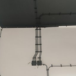 Michal Czuber - Ściana z poprowadzonymi przewodami elektrycznymi w czarnych rurkach, przymocowanymi uchwytami. Widoczne puszki instalacyjne i fragment sufitu w ciemnym kolorze.