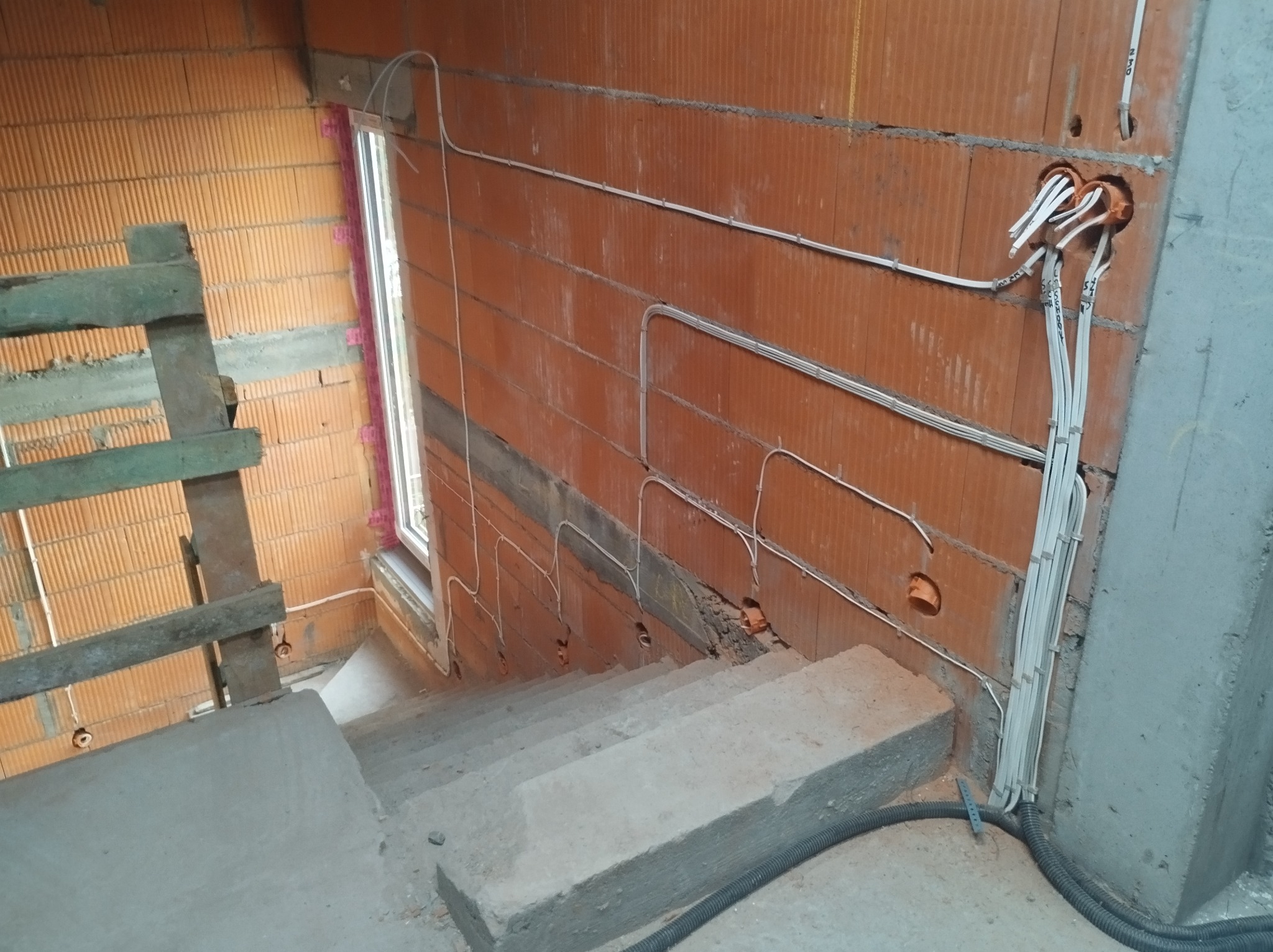 Instalacja elektryczna w surowym budynku: przewody prowadzone po ceglanej ścianie wzdłuż schodów, widoczne puszki instalacyjne i kable. Budynek w trakcie budowy.