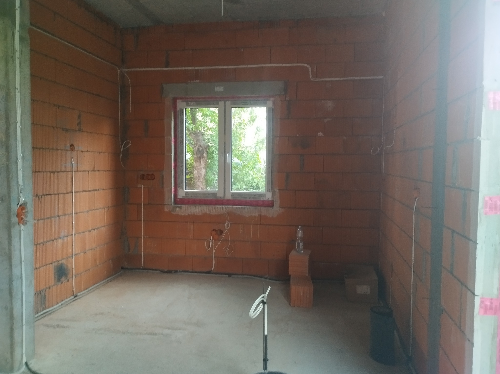 Surowy stan wnętrza z cegły z poprowadzoną instalacją elektryczną w białych rurkach. Widoczne puszki elektryczne i przewody. Okno z widokiem na zieleń.