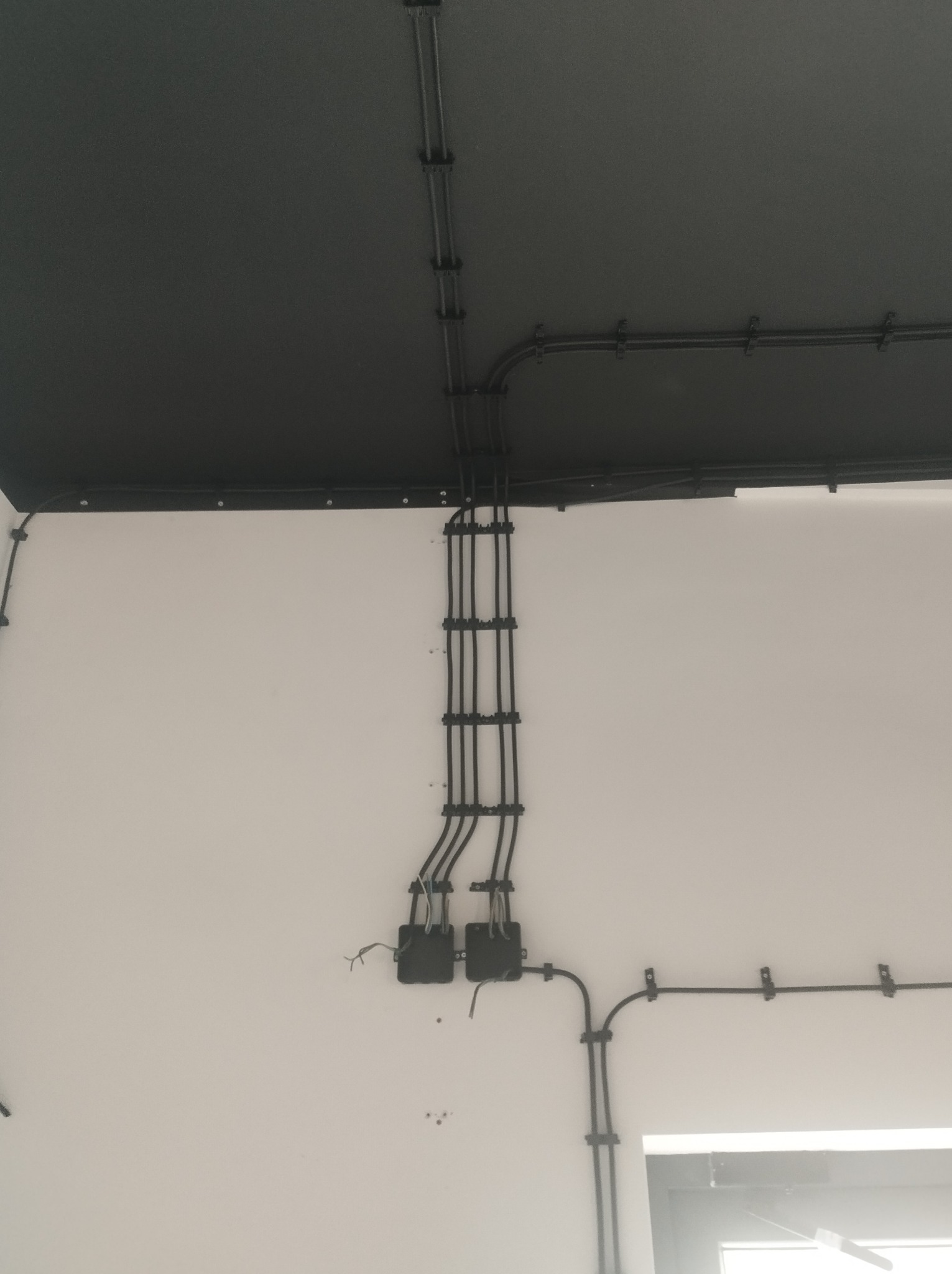 Ściana z poprowadzonymi przewodami elektrycznymi w czarnych rurkach, przymocowanymi uchwytami. Widoczne puszki instalacyjne i fragment sufitu w ciemnym kolorze.