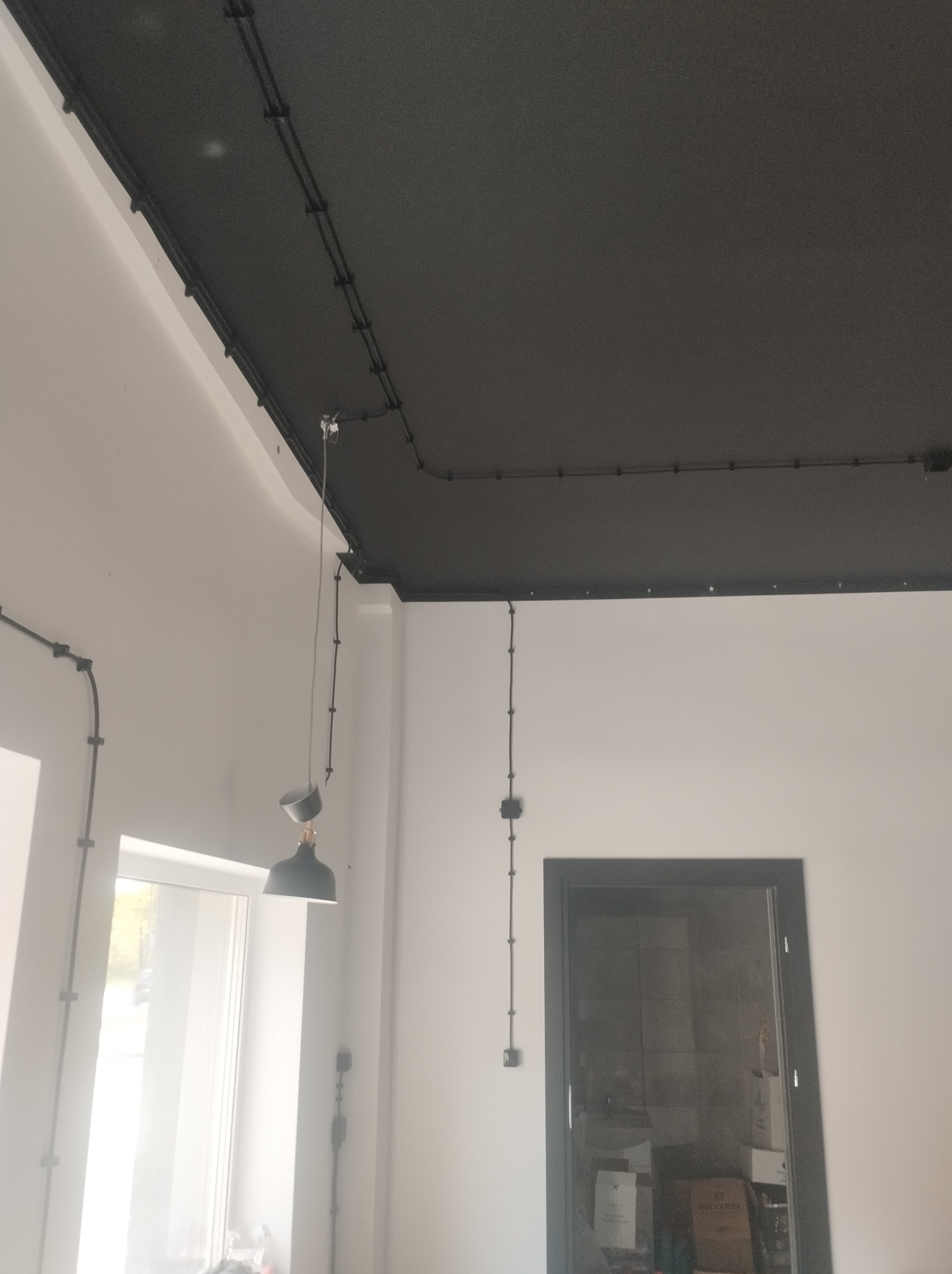 Nowoczesne instalacje elektryczne natynkowe w czarnych peszlach na suficie i ścianach, minimalistyczny design z lampą wiszącą. Widok na okno i drzwi.